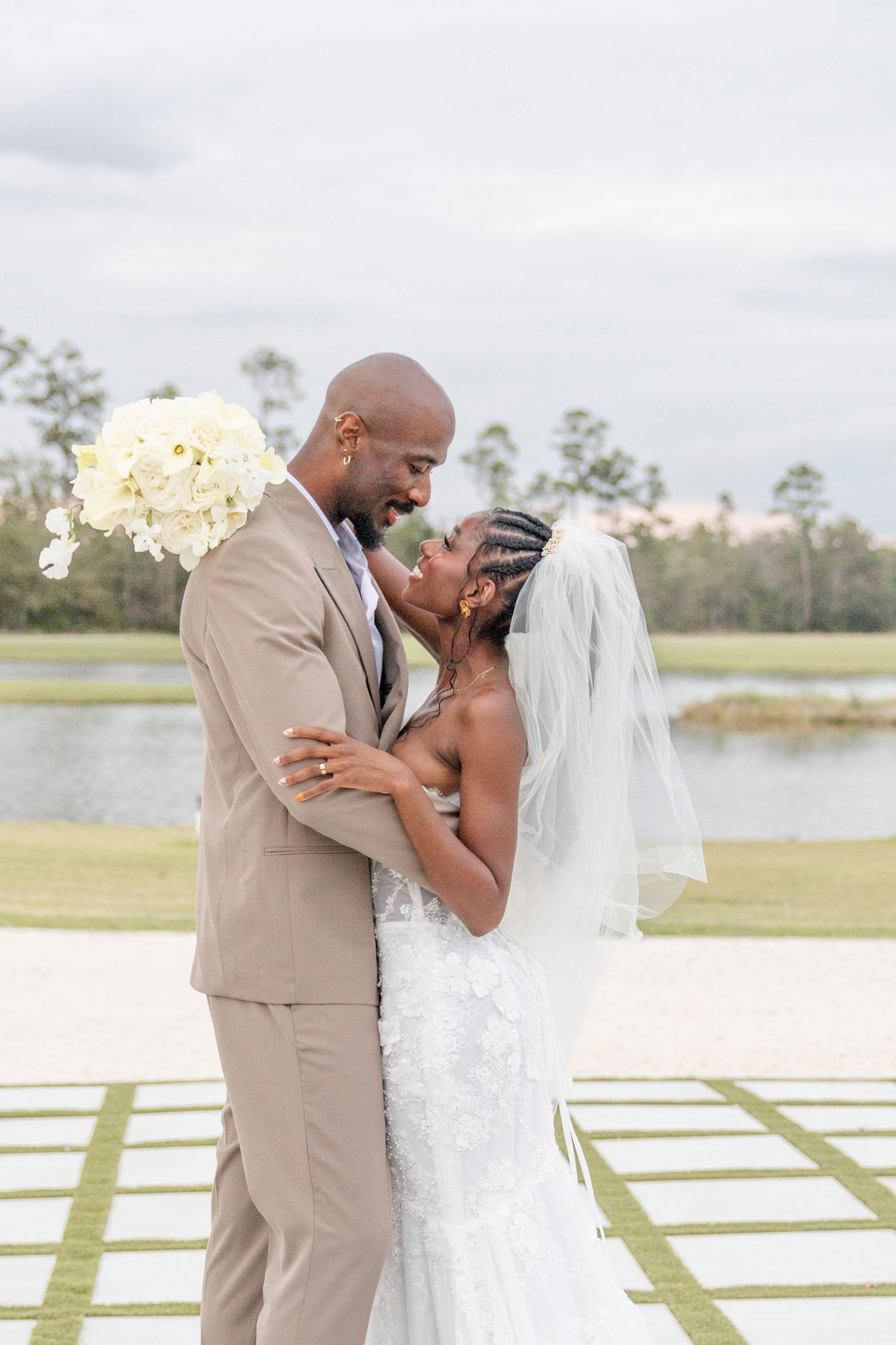 A couple share a tender moment in wedding attire on a patio overlooking a lake, with the bride holding white flowers.