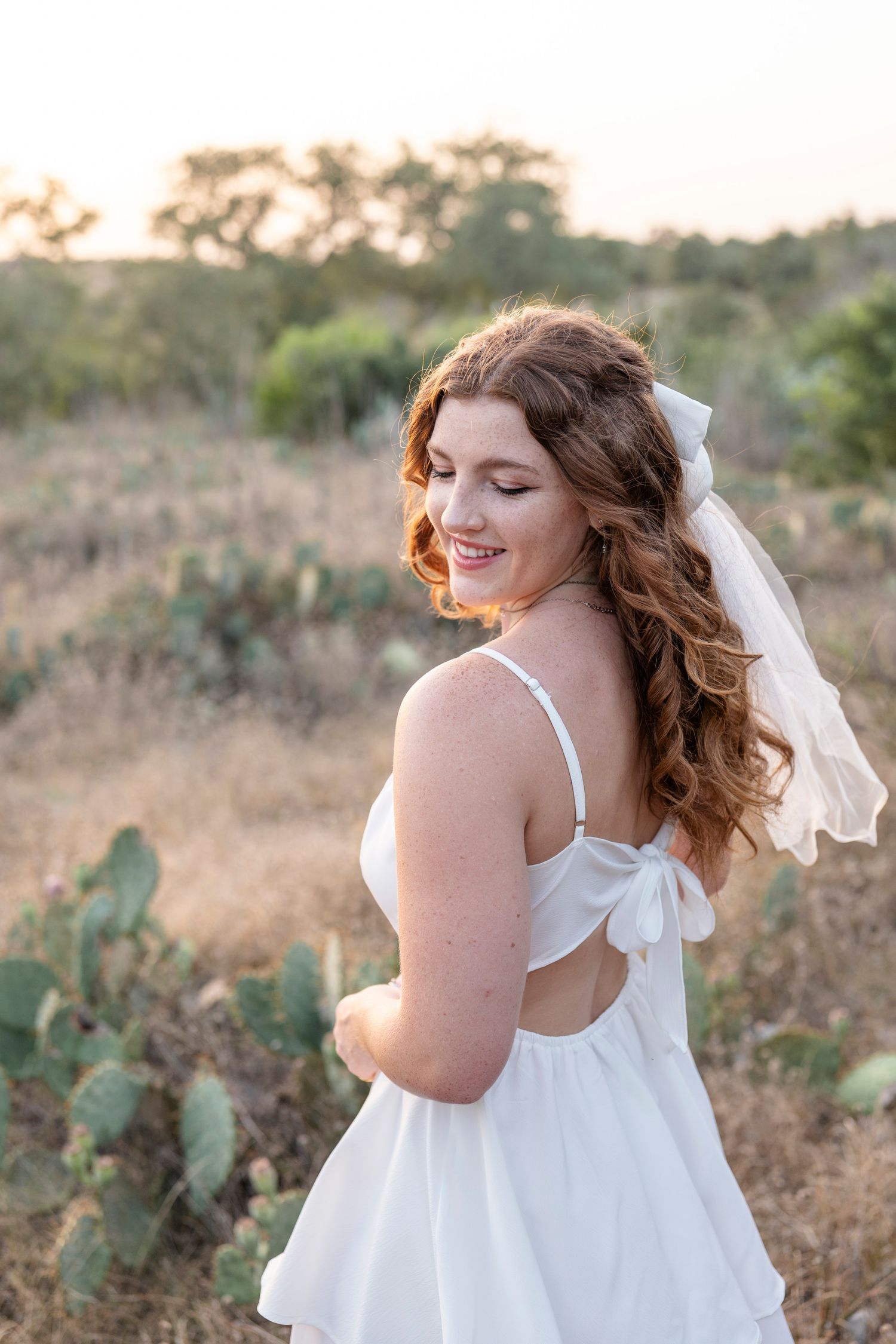 A bride in a flowing white dress poses in a desert landscape with prickly pear cactus at golden hour.