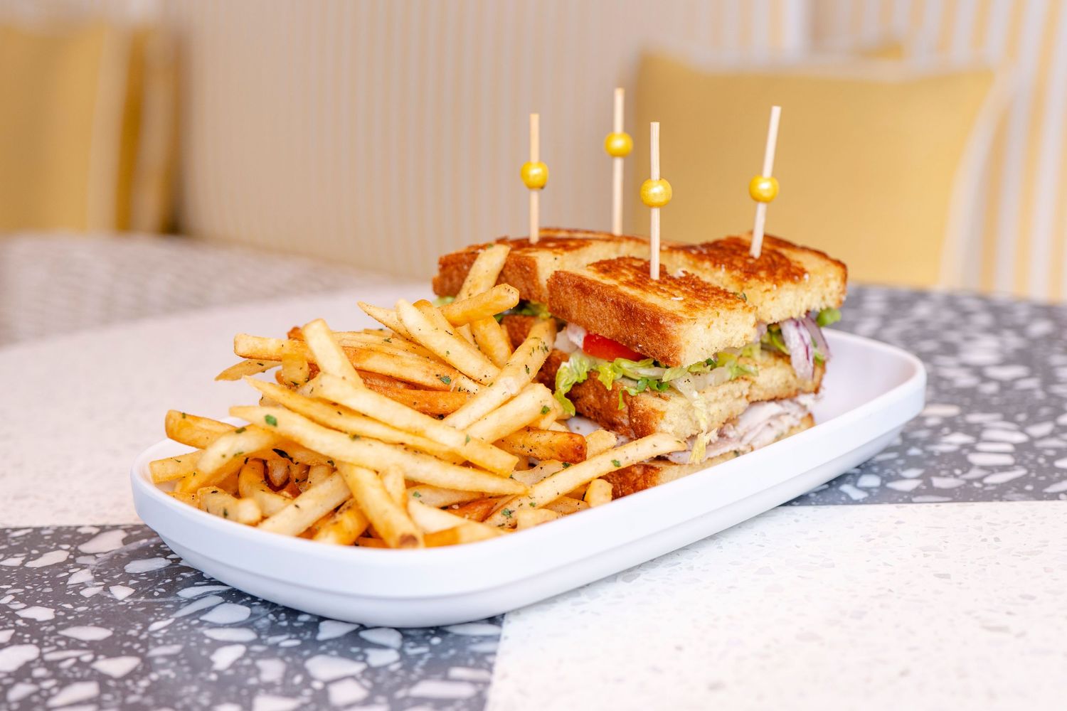 Club sandwich served on a white plate with golden french fries and toothpicks on a granite counter.