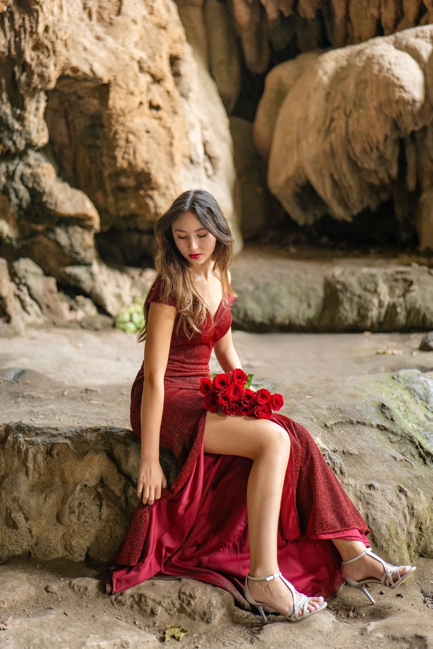 Woman in flowing red evening gown and heels poses against rocky cave backdrop with red roses.