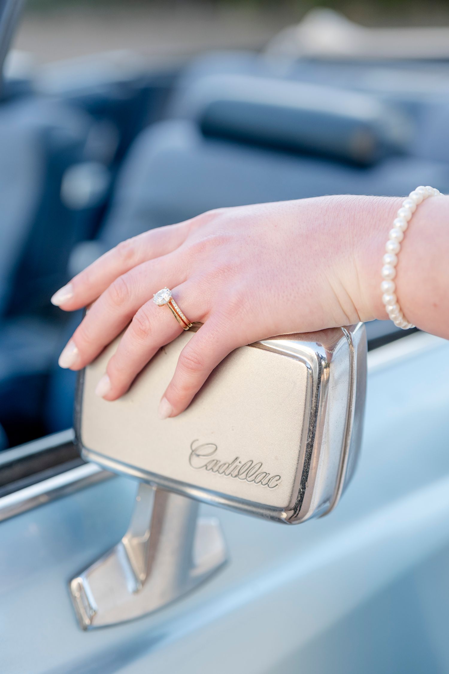A delicate pearl bracelet and wedding ring adorned hand rests on a vintage Cadillac side mirror.