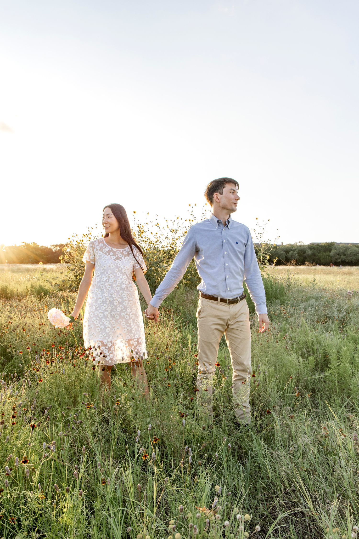 Two people walk hand in hand through a wildflower meadow at sunset while sharing a romantic moment in nature.