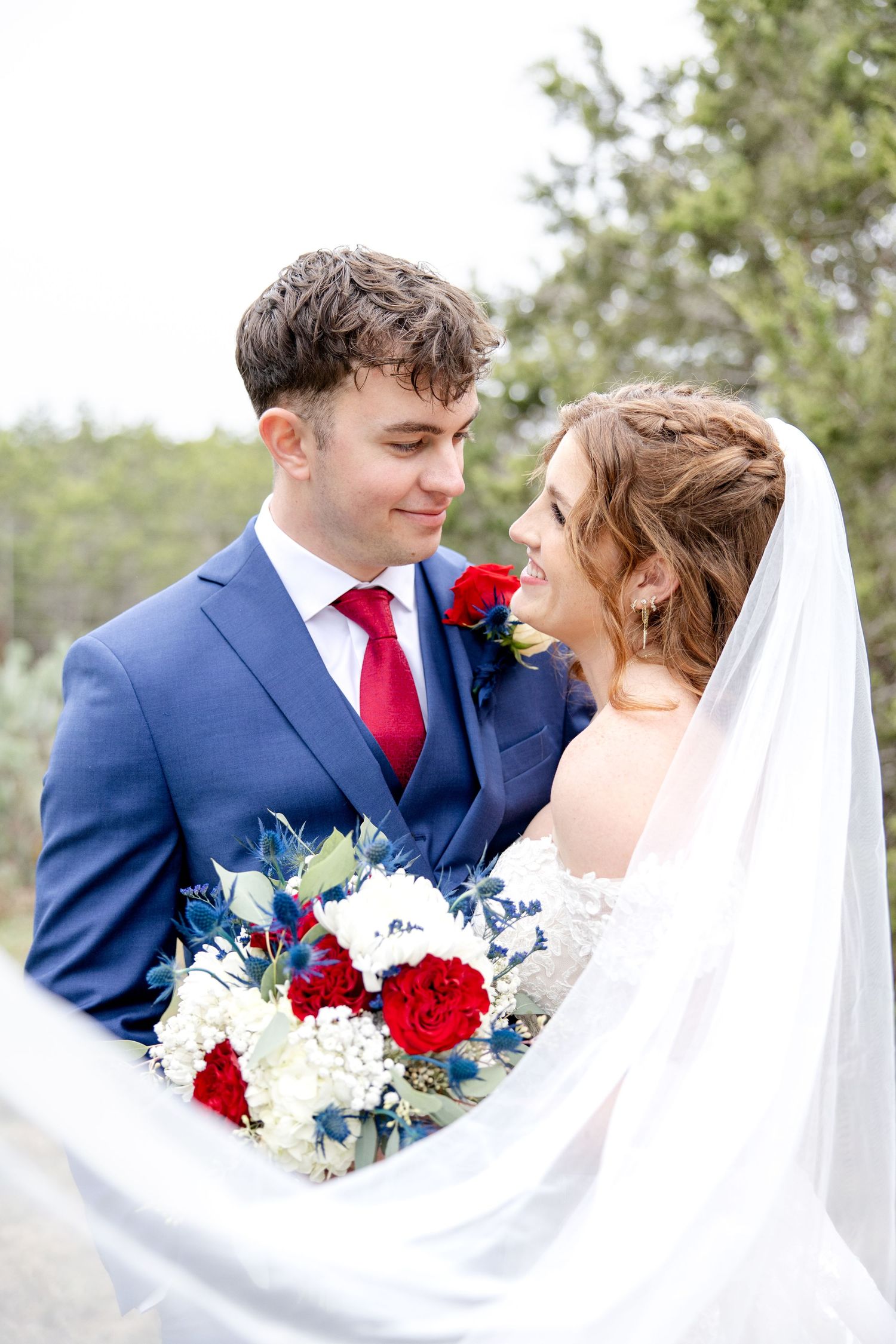 A bride and groom share an intimate moment surrounded by foliage, with red and white floral bouquet and flowing veil.
