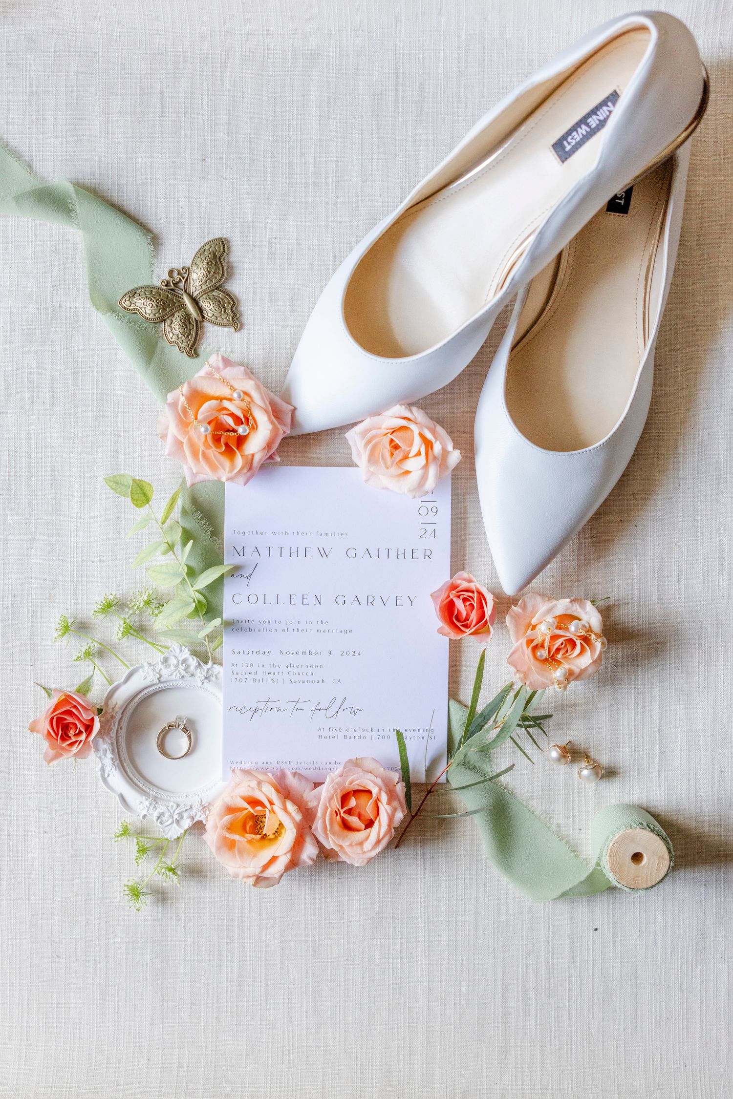 Elegant white bridal shoes arranged with peach roses and wedding invitation on light fabric background.