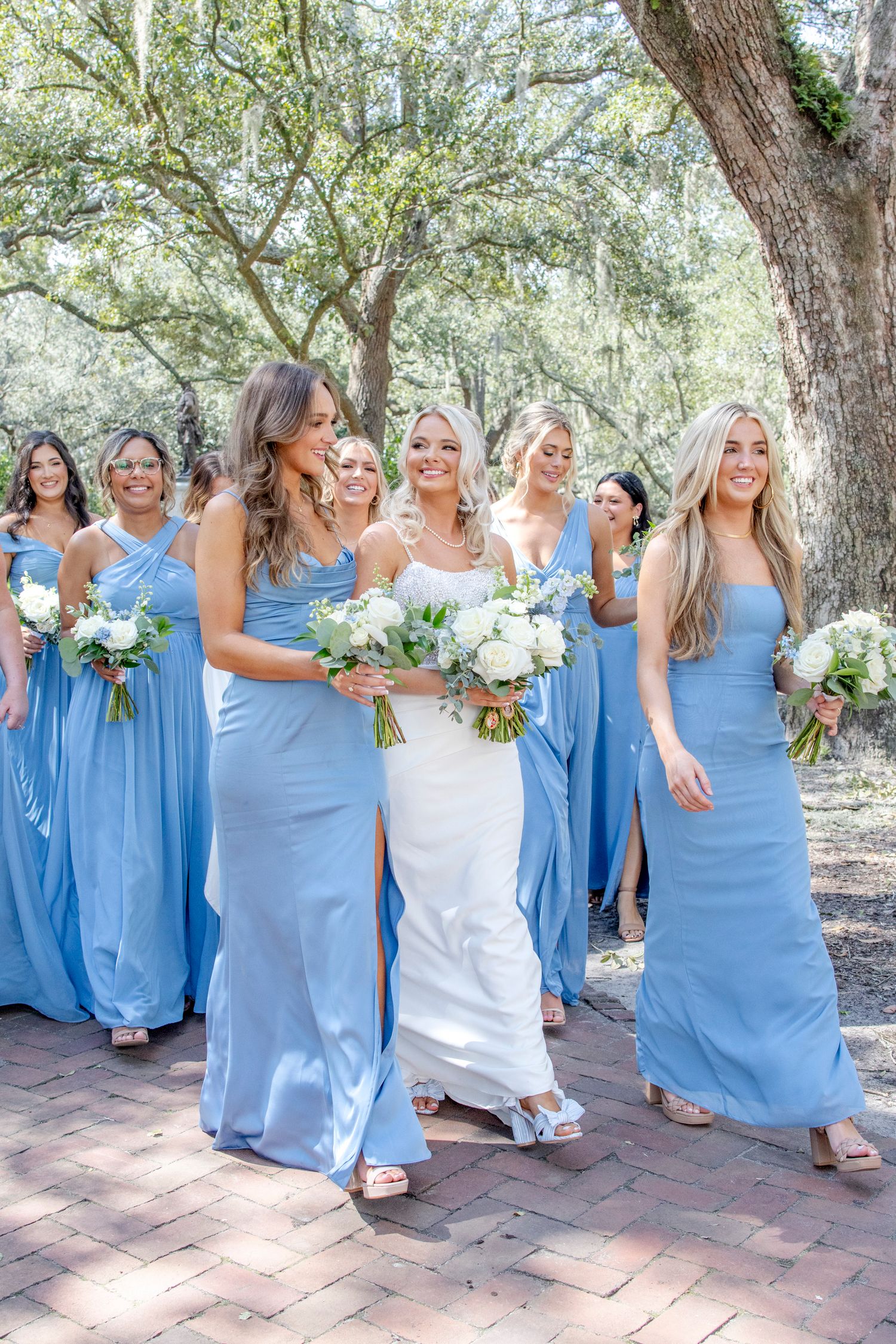 Bridesmaids in flowing light blue dresses carrying white bouquets walk together outdoors among trees on a brick path.