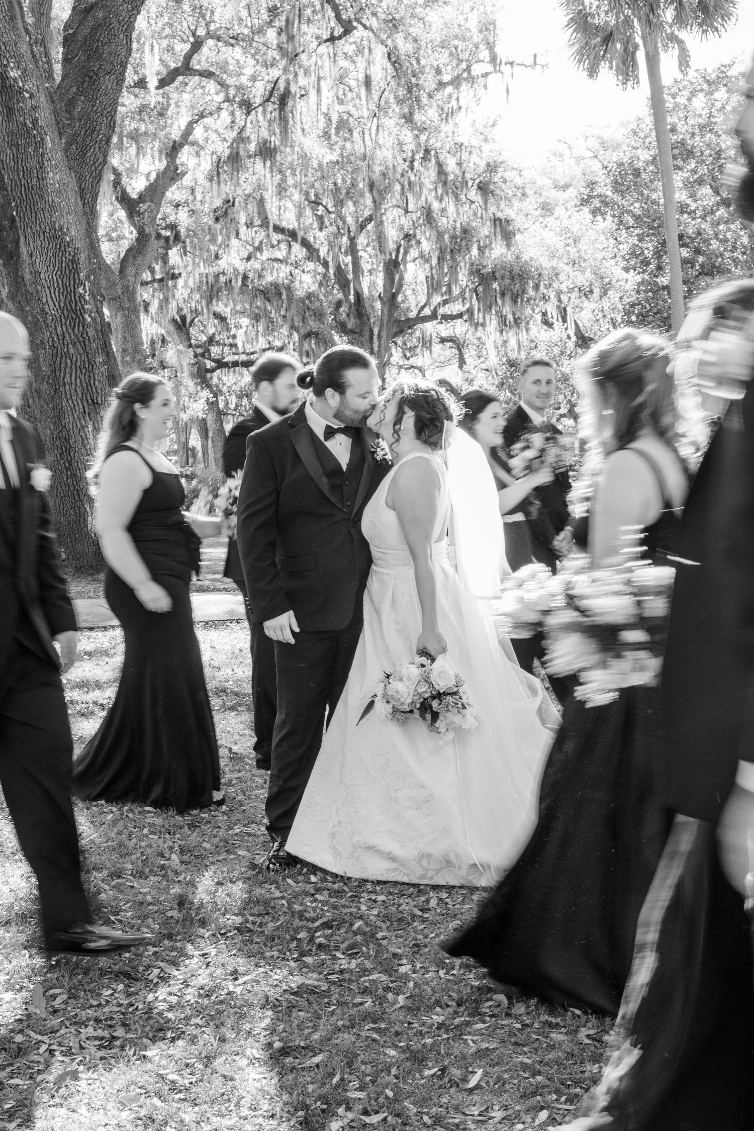 A wedding party gathers under Spanish moss-draped trees in a black and white outdoor photograph.