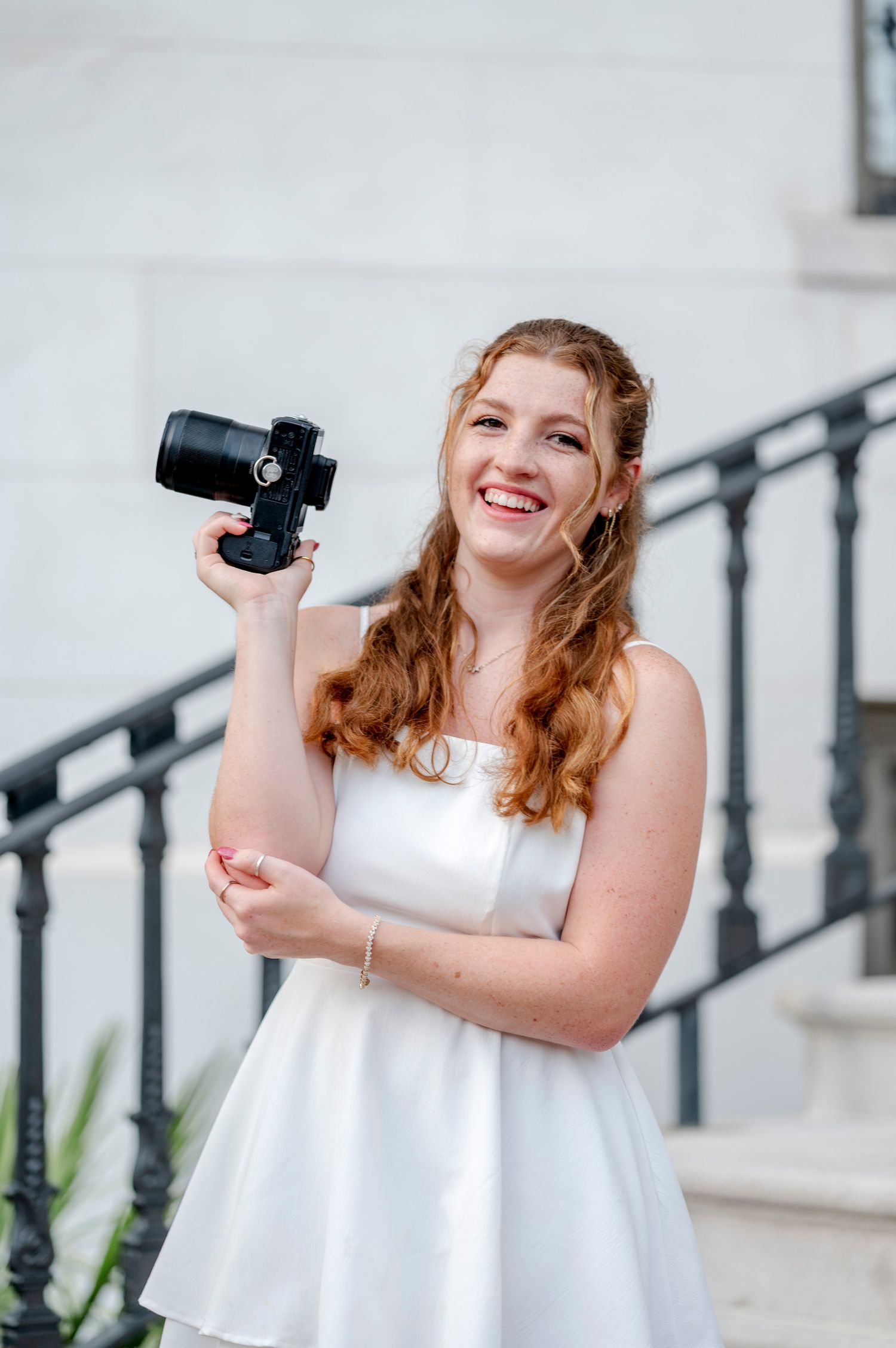 A person in white curved top smiles while holding a camera against a light background in professional photos.