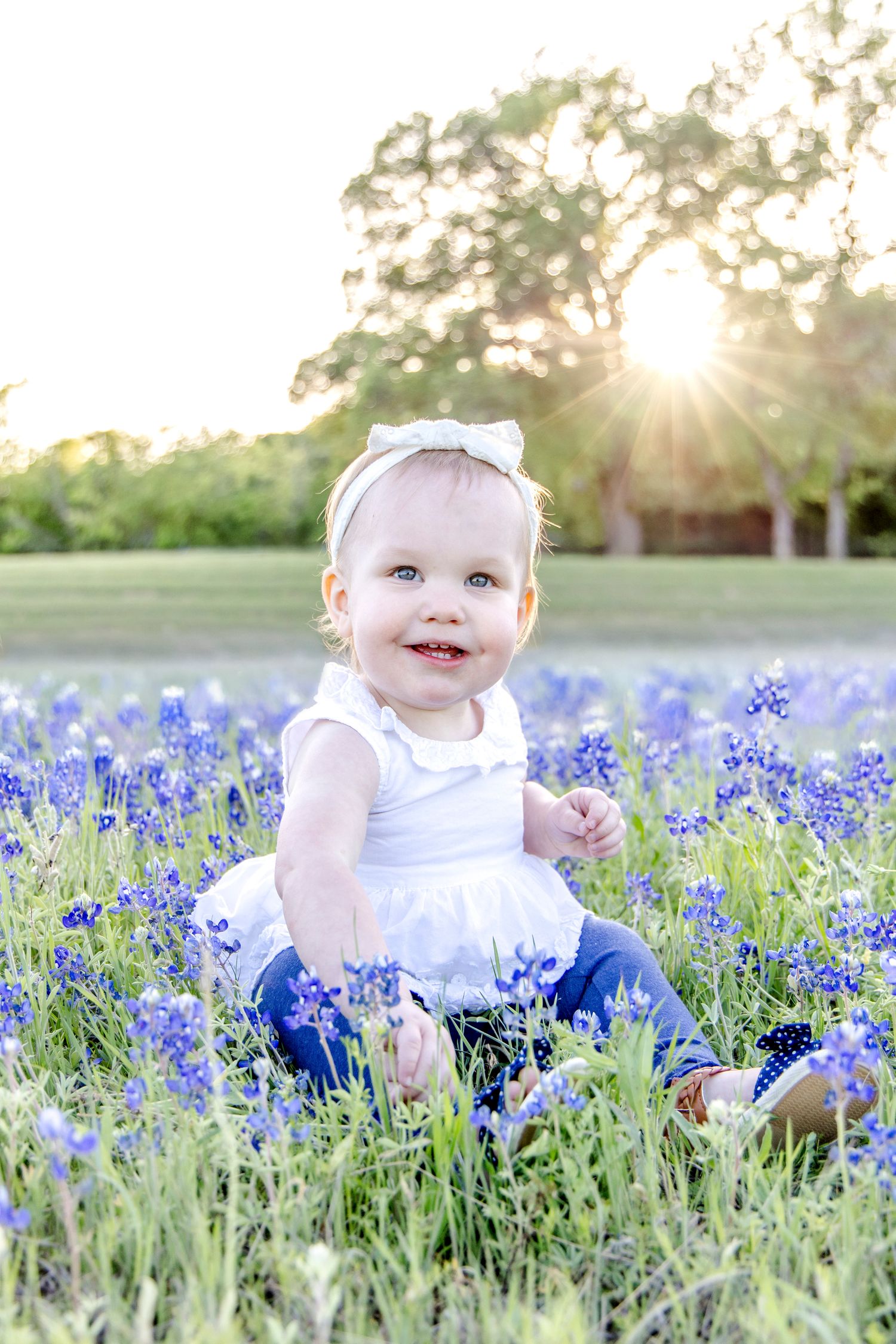 A small child in a white dress sits among blooming bluebonnets at sunset in a grassy field.