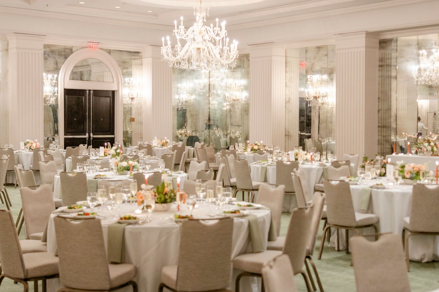 Elegant ballroom with crystal chandeliers and round tables set for a formal wedding reception in white and cream decor.