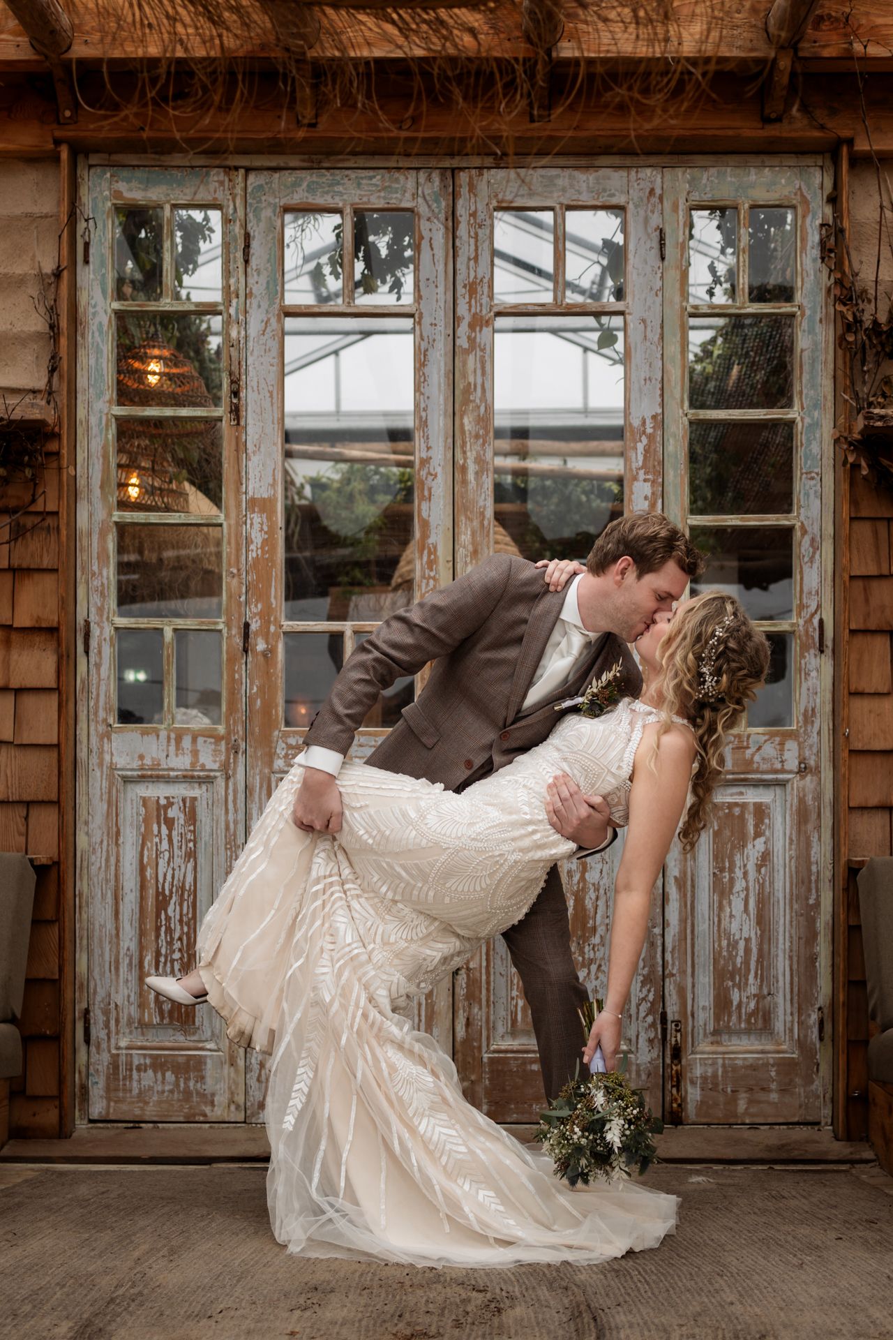 Romantic wedding couple shares a passionate dip kiss in front of rustic wooden French doors with distressed paint.