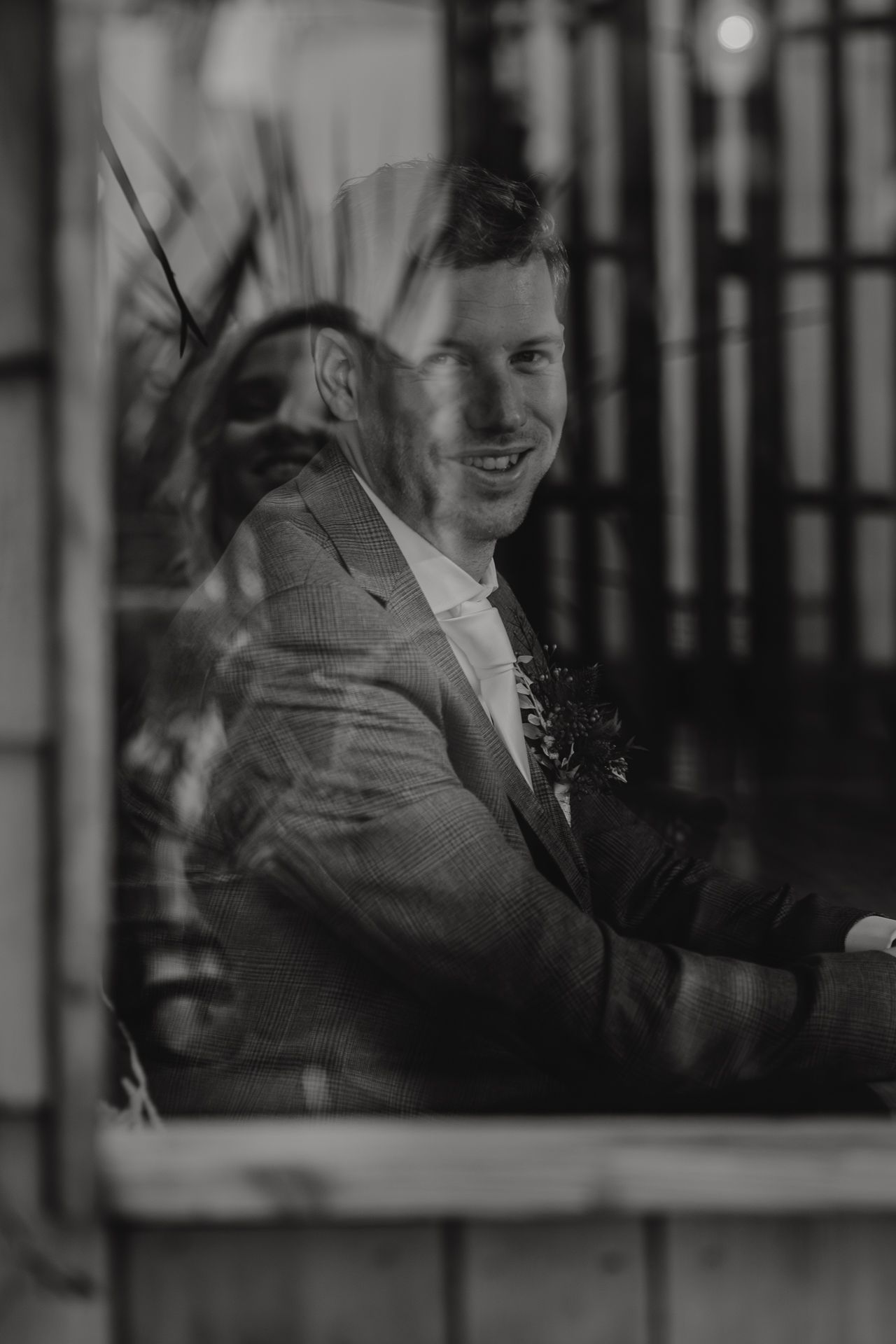 Black and white portrait of a groom in a suit with boutonniere smiling through a window at a wedding venue.