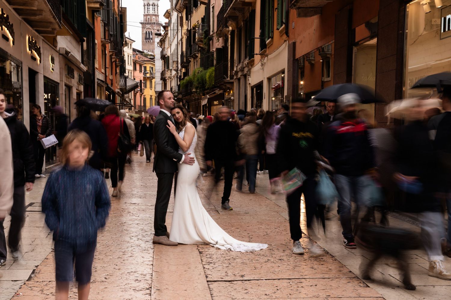 A couple in wedding attire standing amid a bustling street filled with motion-blurred pedestrians in a historic Italian town.