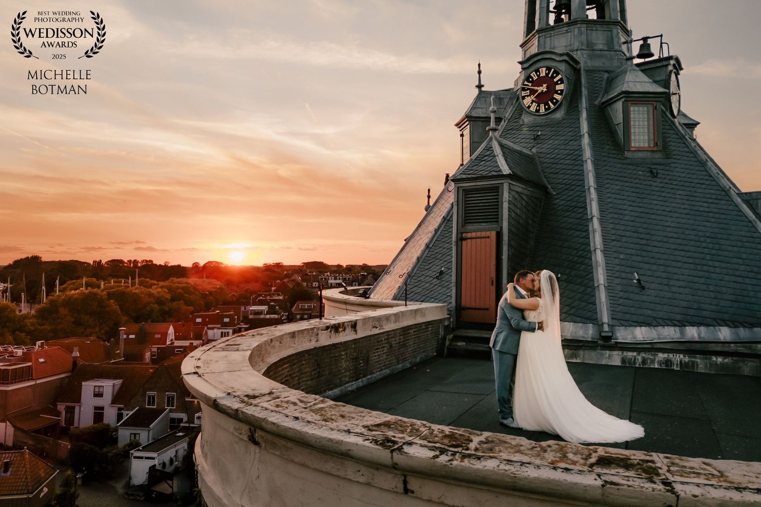 Couple in wedding attire poses on historic lighthouse balcony during dramatic orange sunset along coastal shoreline.