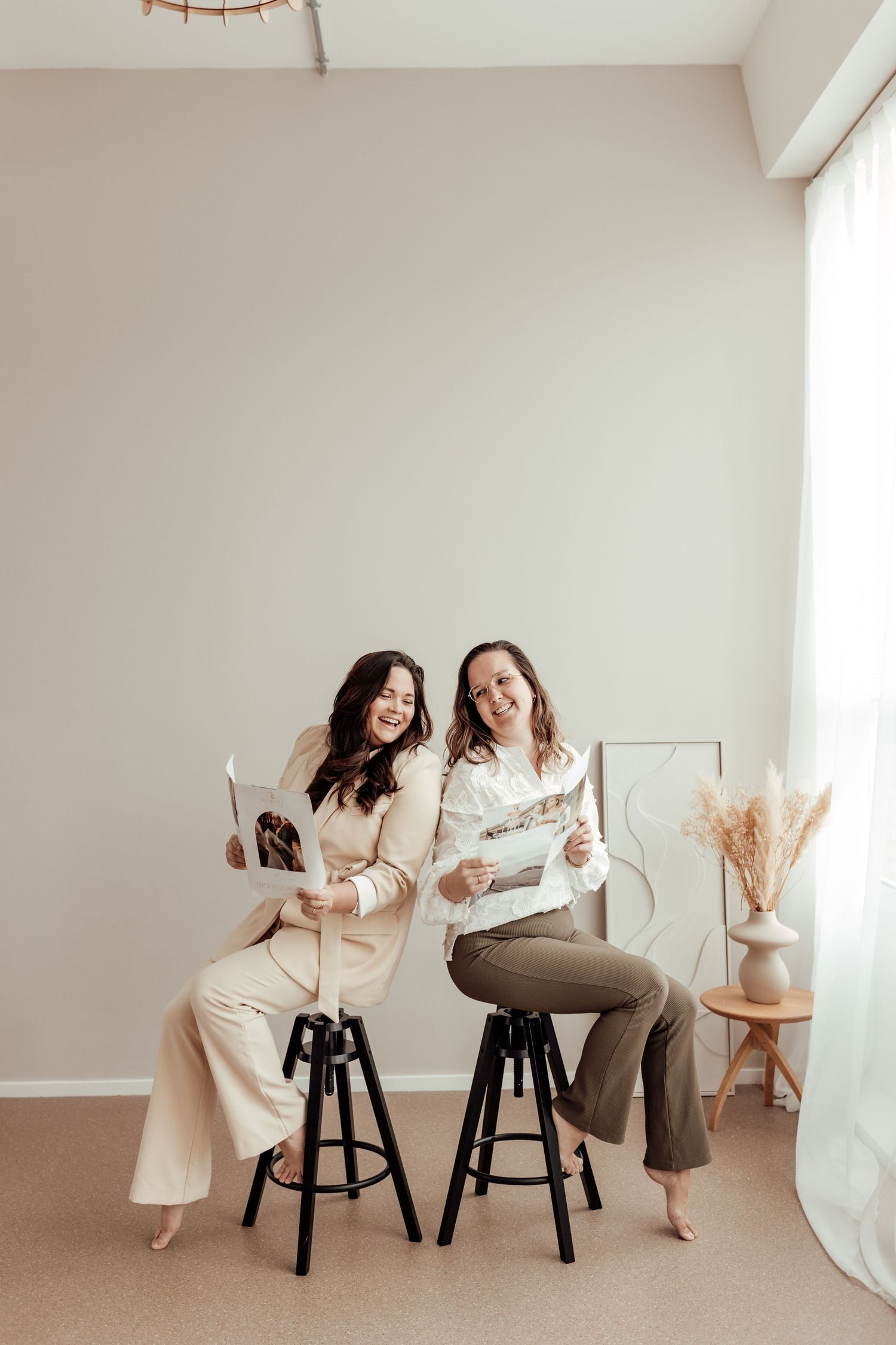 Two women sitting on black bar stools share a laugh while holding papers in a minimalist room with pampas grass decor.