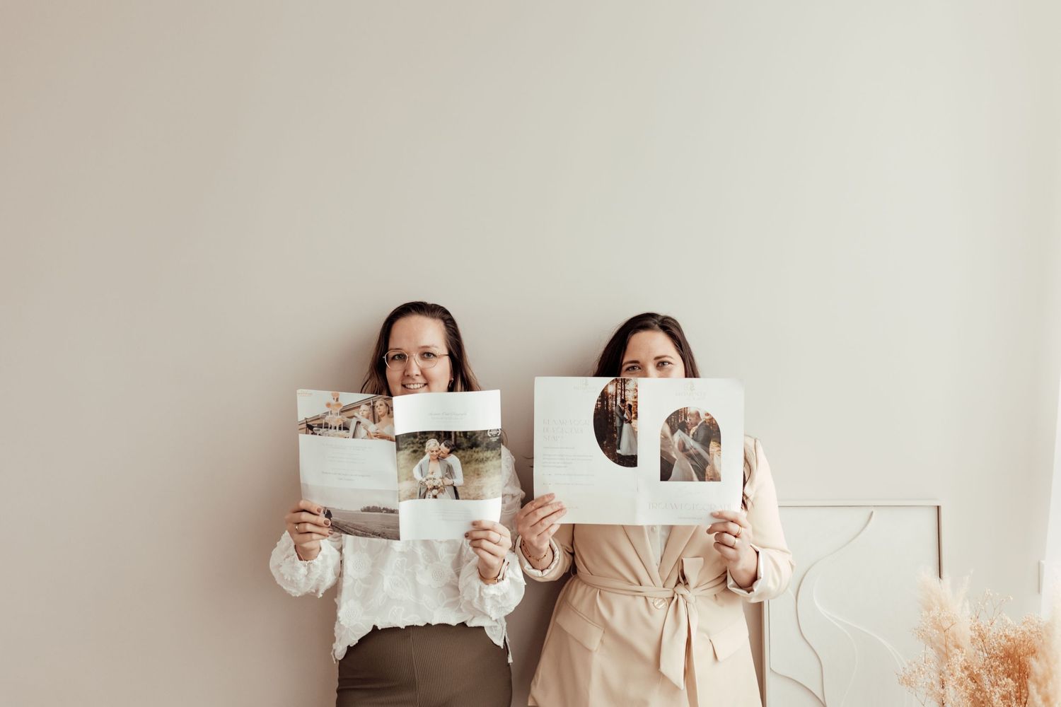Two people hold up wedding photos while posing against a neutral wall with dried pampas grass decor.