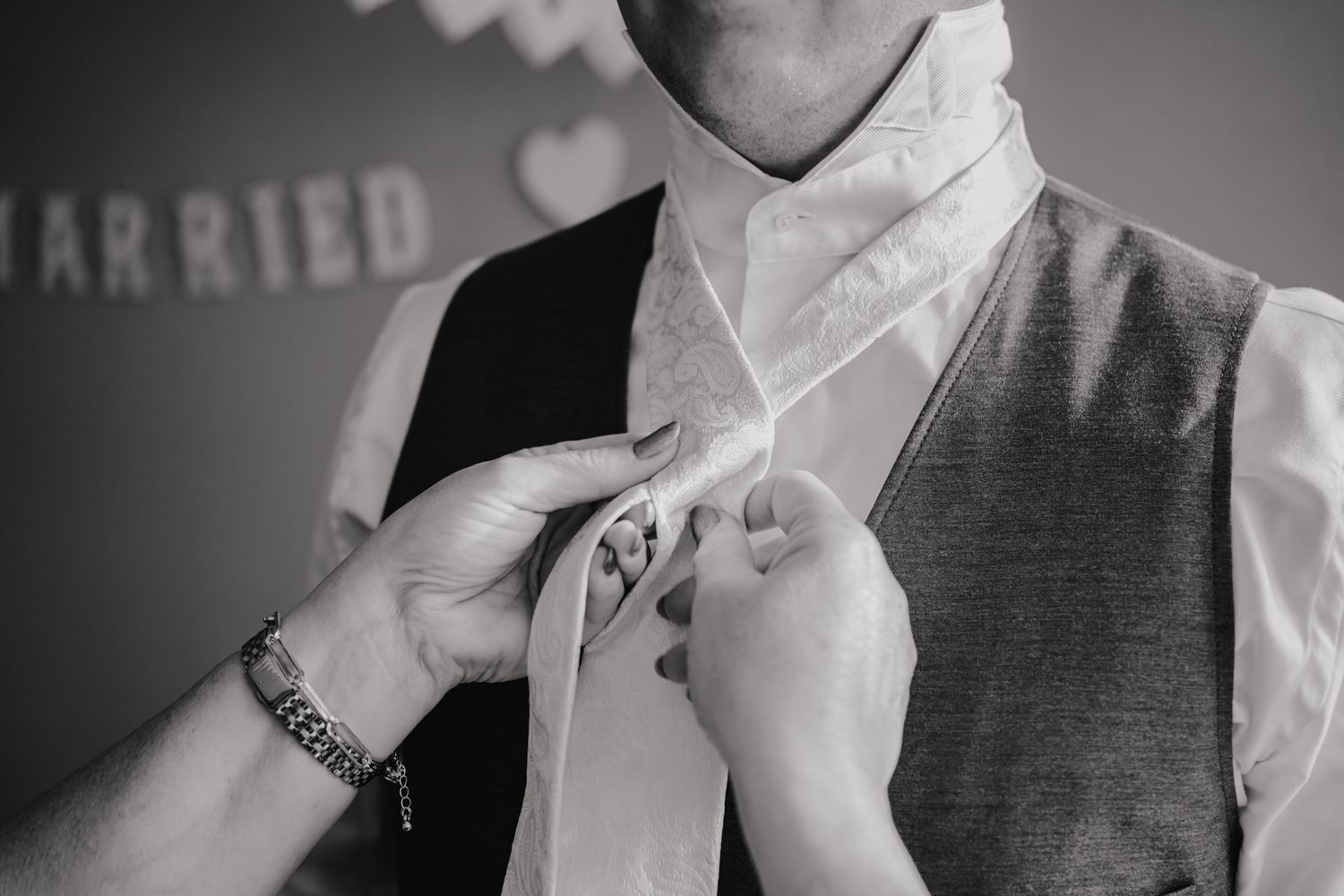 Helper adjusts groom's tie in black and white wedding preparation photo showing hands at neckline and formal vest.