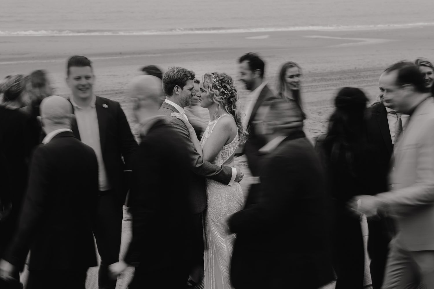 A black and white artistic photo of a wedding couple embracing surrounded by guests at an outdoor ceremony.