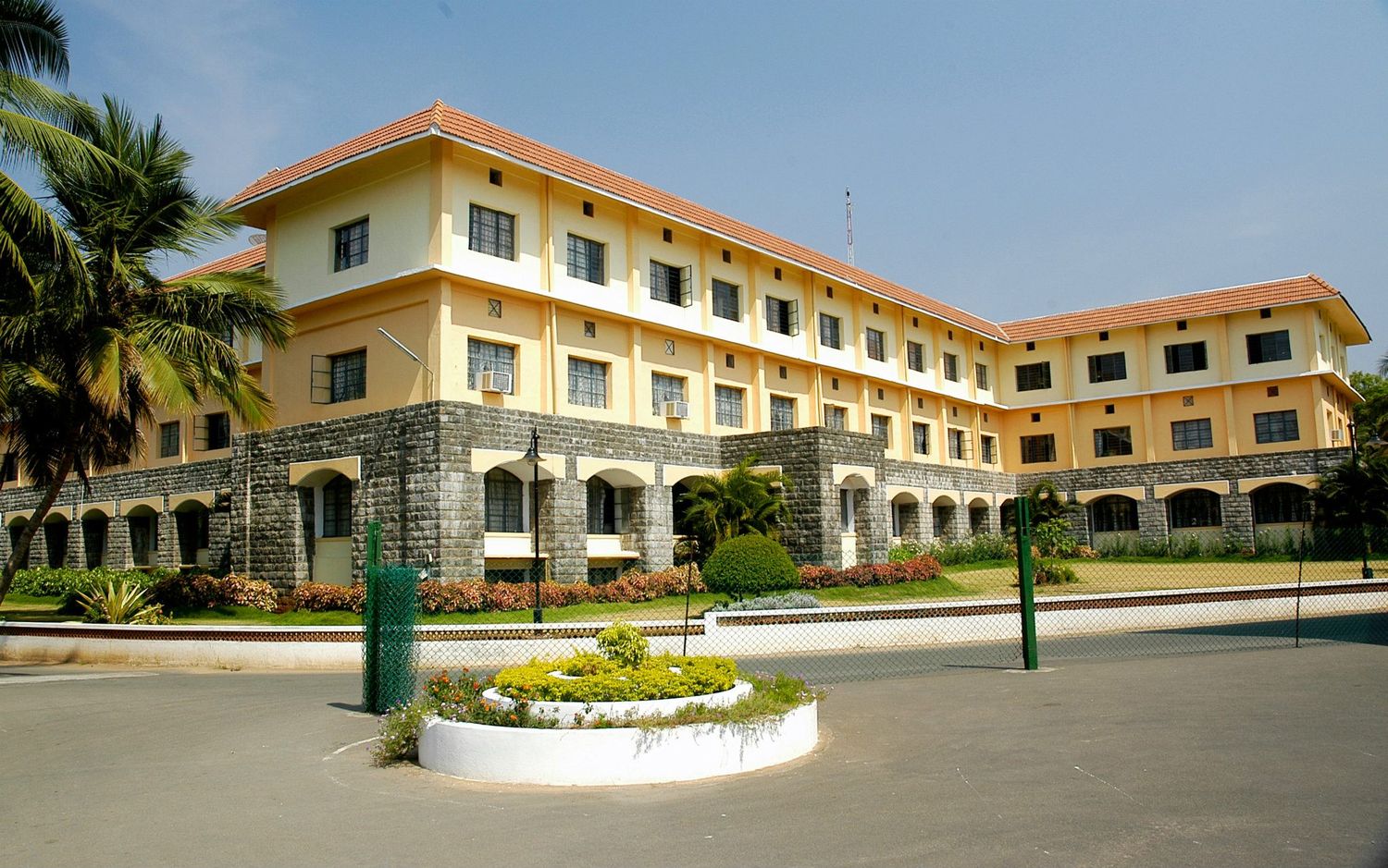 Yellow colonial building with palm trees and circular garden feature in front.
