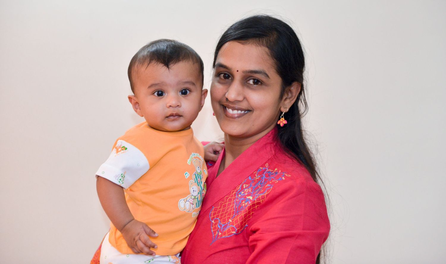 A parent wearing a bright red top holds a baby dressed in an orange outfit against a neutral background.