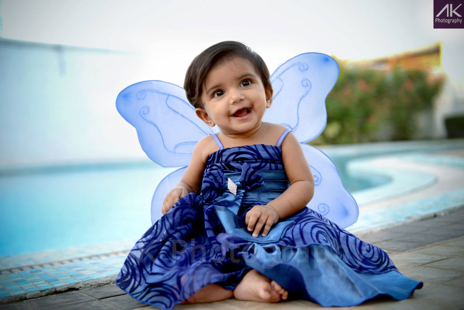 Baby wearing blue dress and butterfly wings sitting by pool.