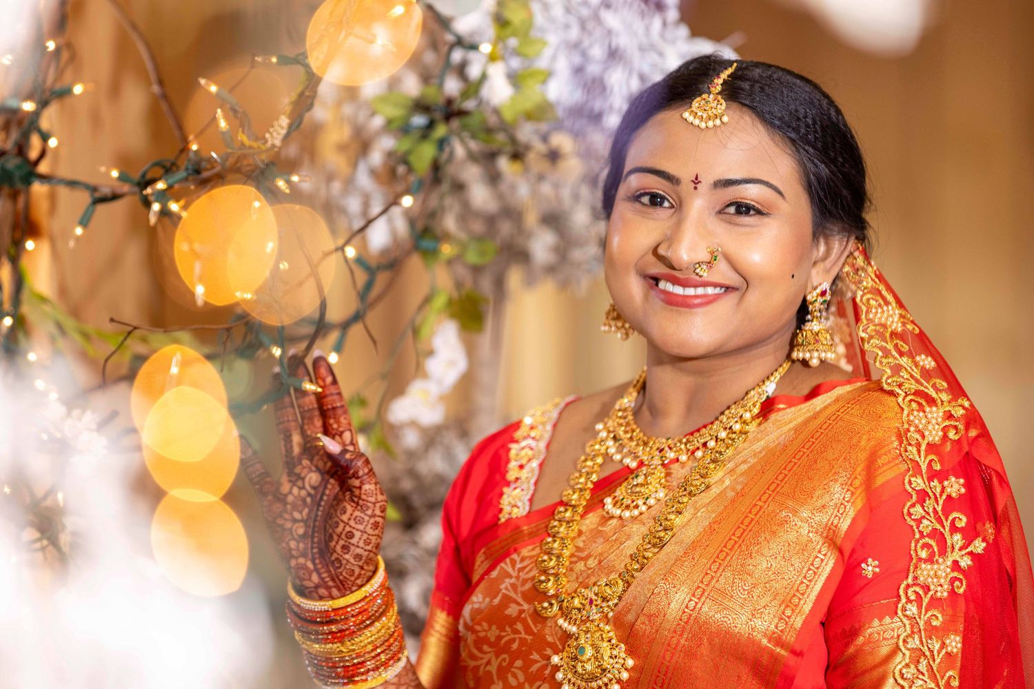 Traditional South Asian bride in red and gold attire posing with decorative lights and flowers.