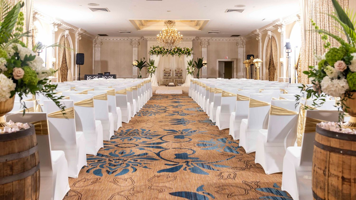 Elegant wedding ceremony aisle with white chairs and floral arch in a luxurious ballroom.