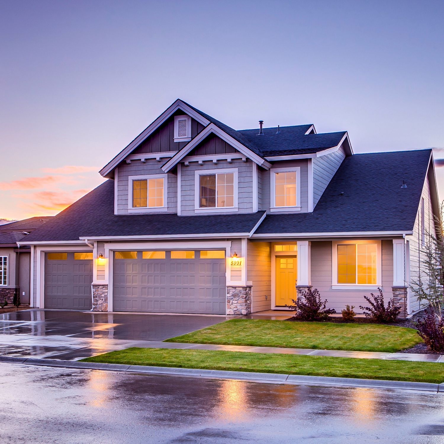 Modern two-story craftsman home with glowing windows and illuminated garage at sunset on wet suburban street.
