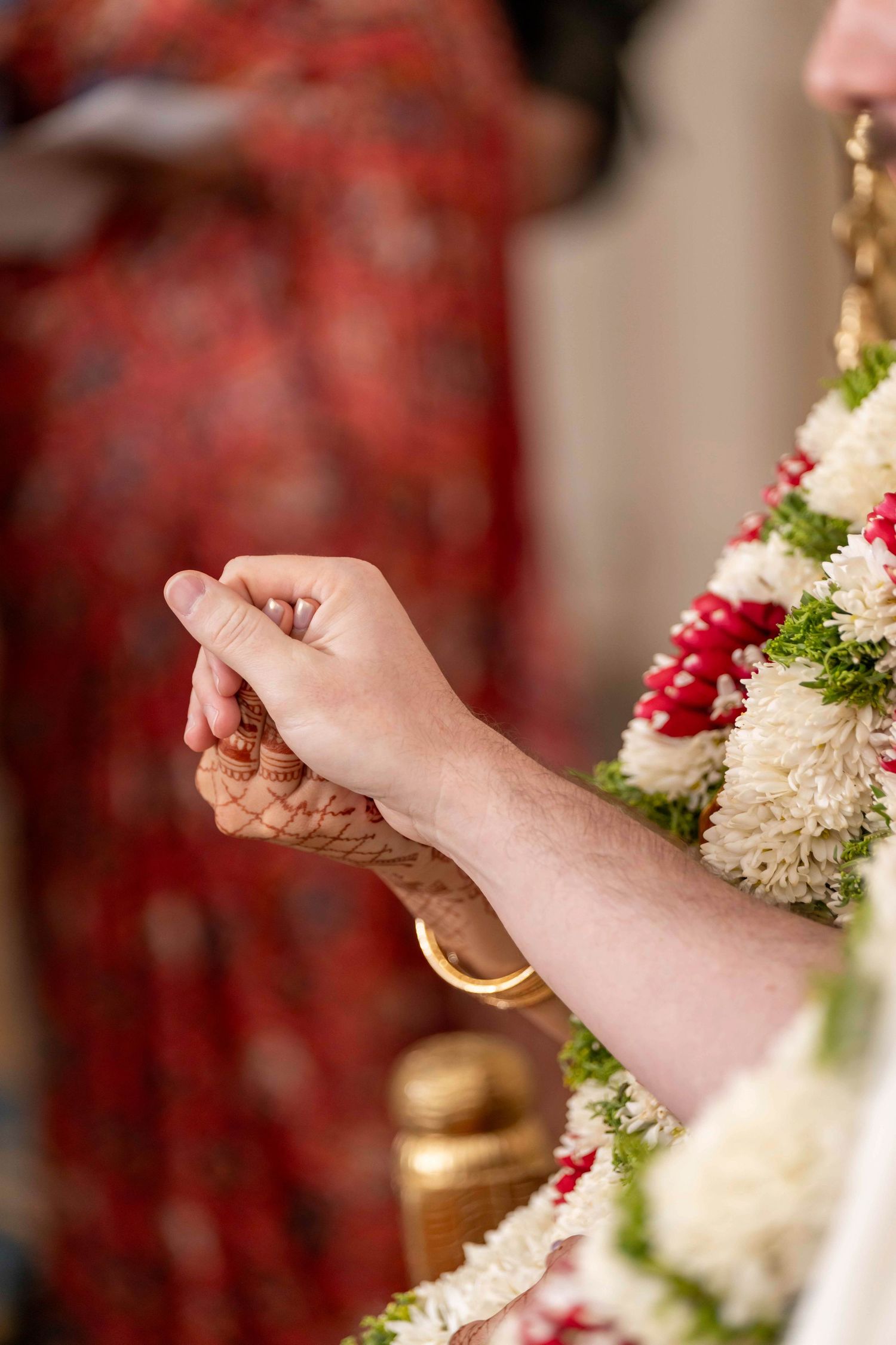 Two people hold hands during a traditional Indian wedding ceremony with floral decorations and gold accents in the background.