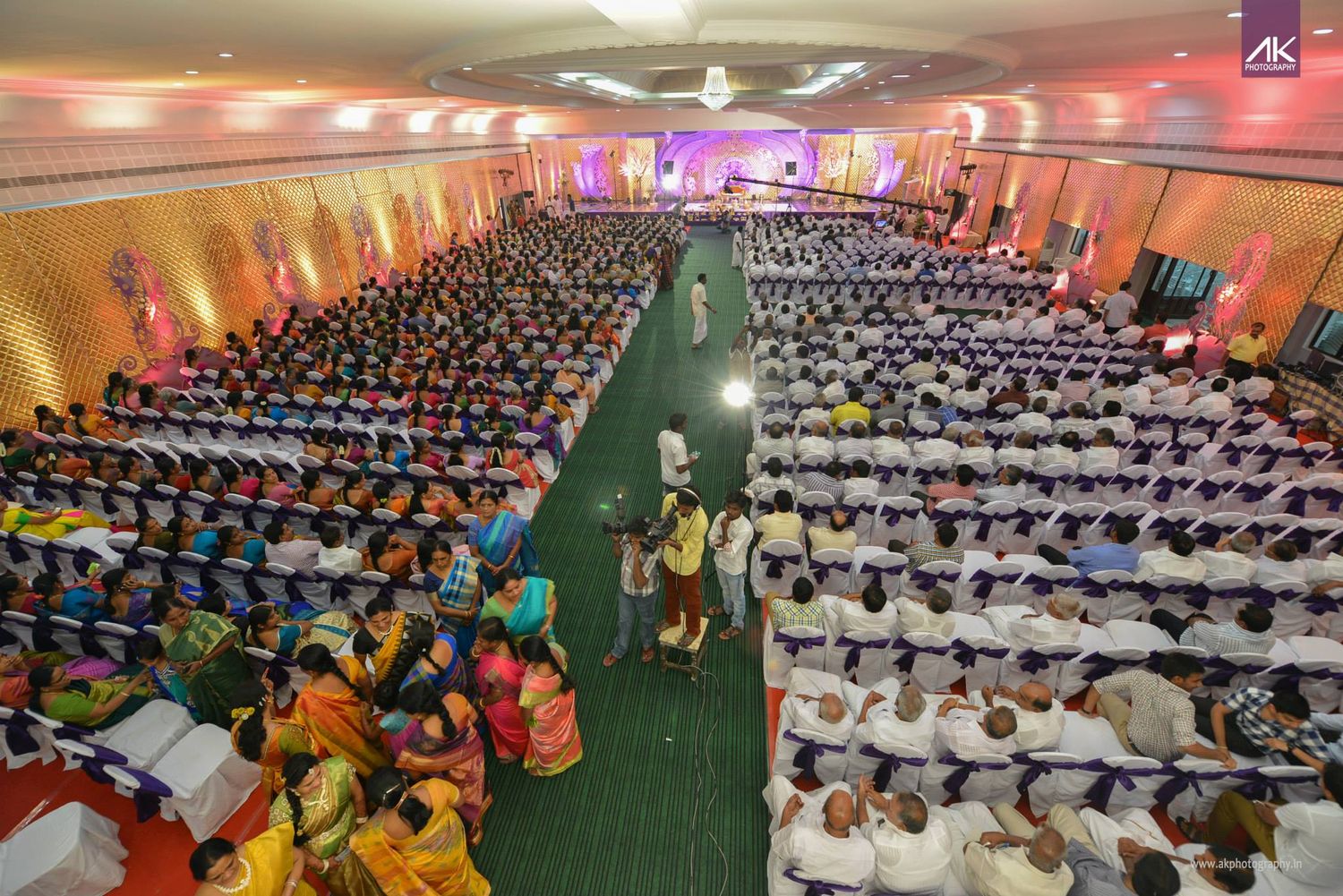Large indoor gathering with rows of people seated in a grand hall with colorful lighting.