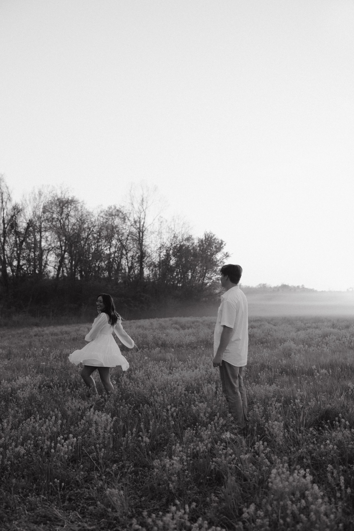 Black and white photo of a couple in a meadow at sunset with trees in the background.