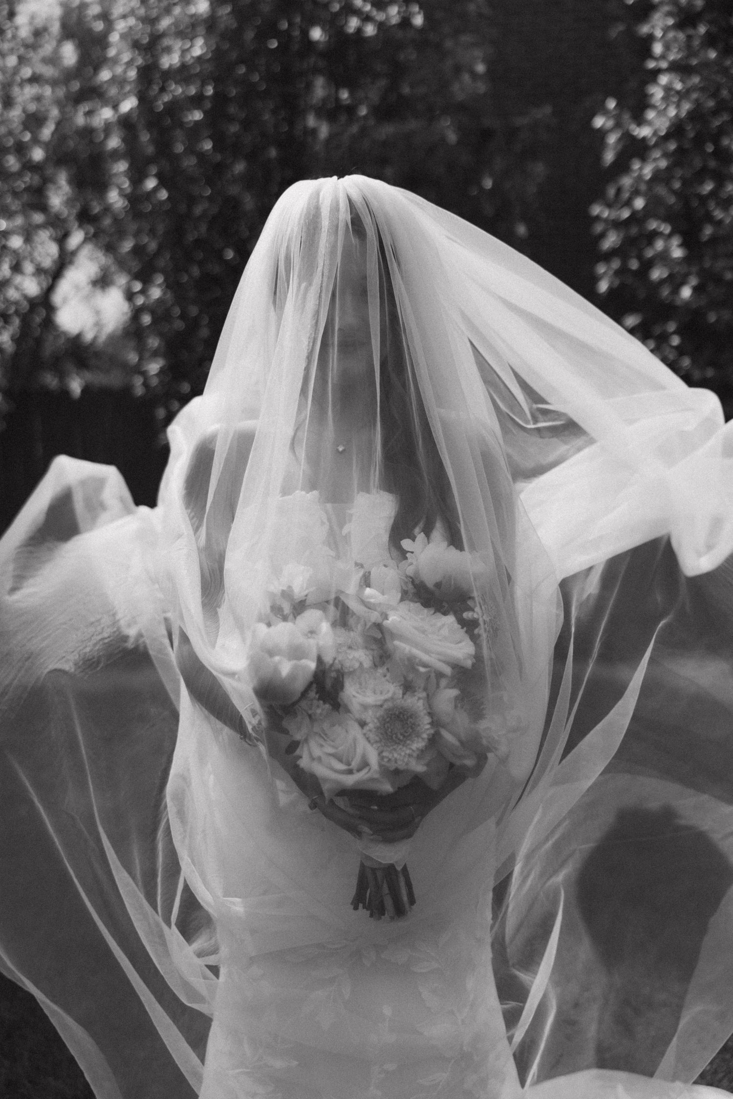 A bride in a flowing veil holds a bouquet of white flowers in this artistic black and white photograph.