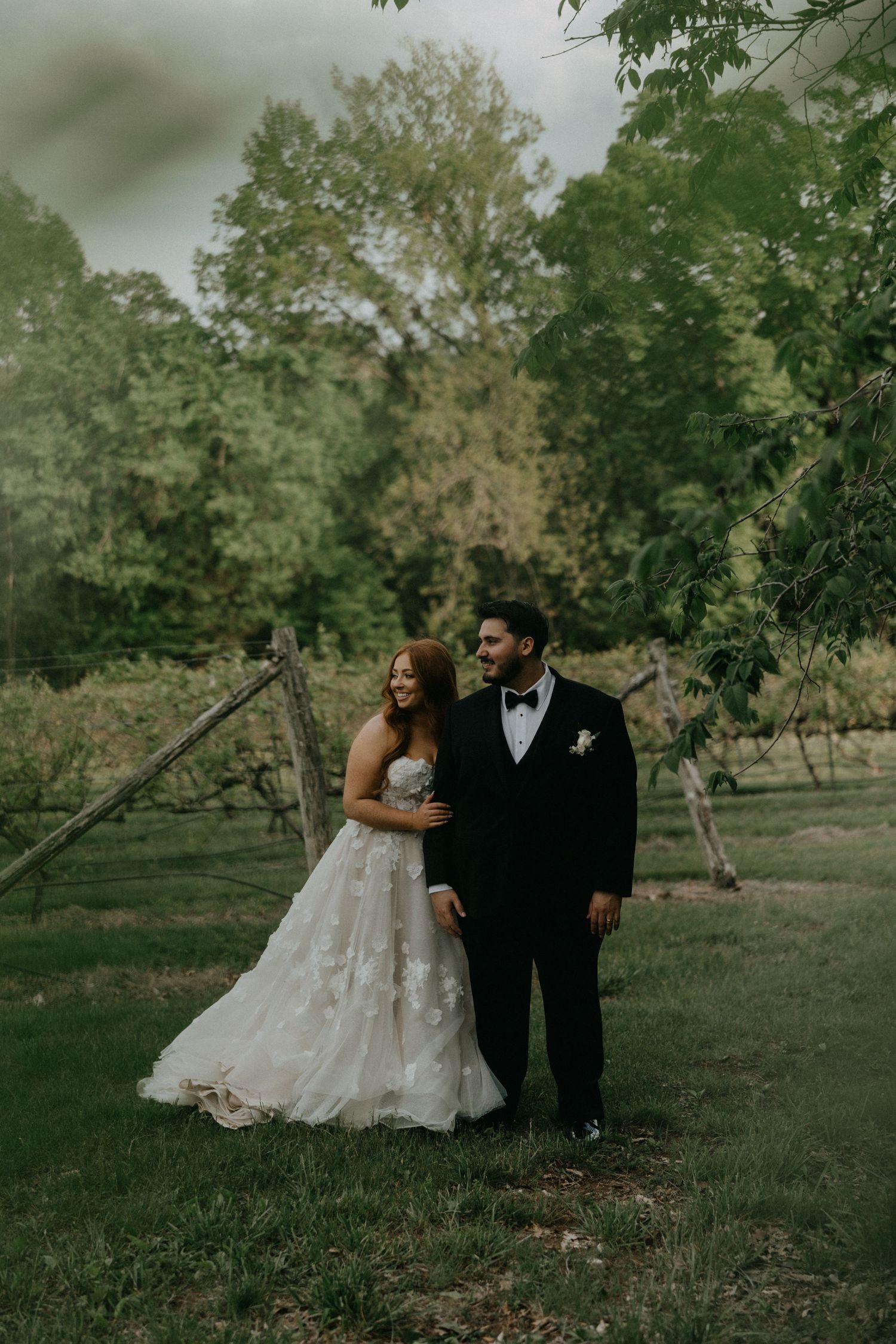 Wedding couple poses candidly among trees in rustic orchard setting.