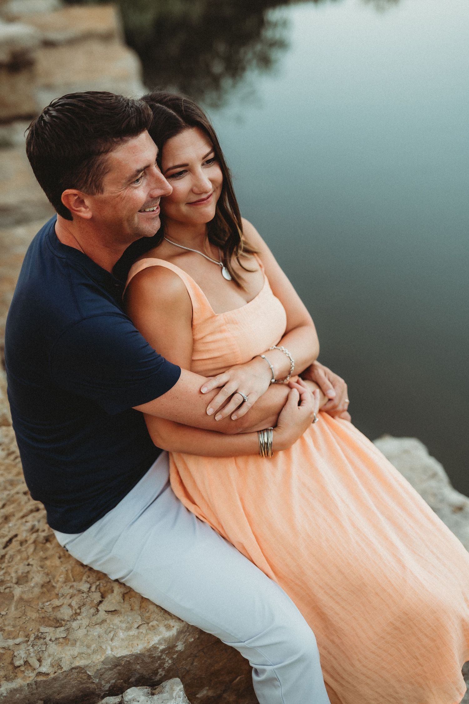 A romantic couple sits by a tranquil lake at sunset, embracing while perched on a rocky shoreline.