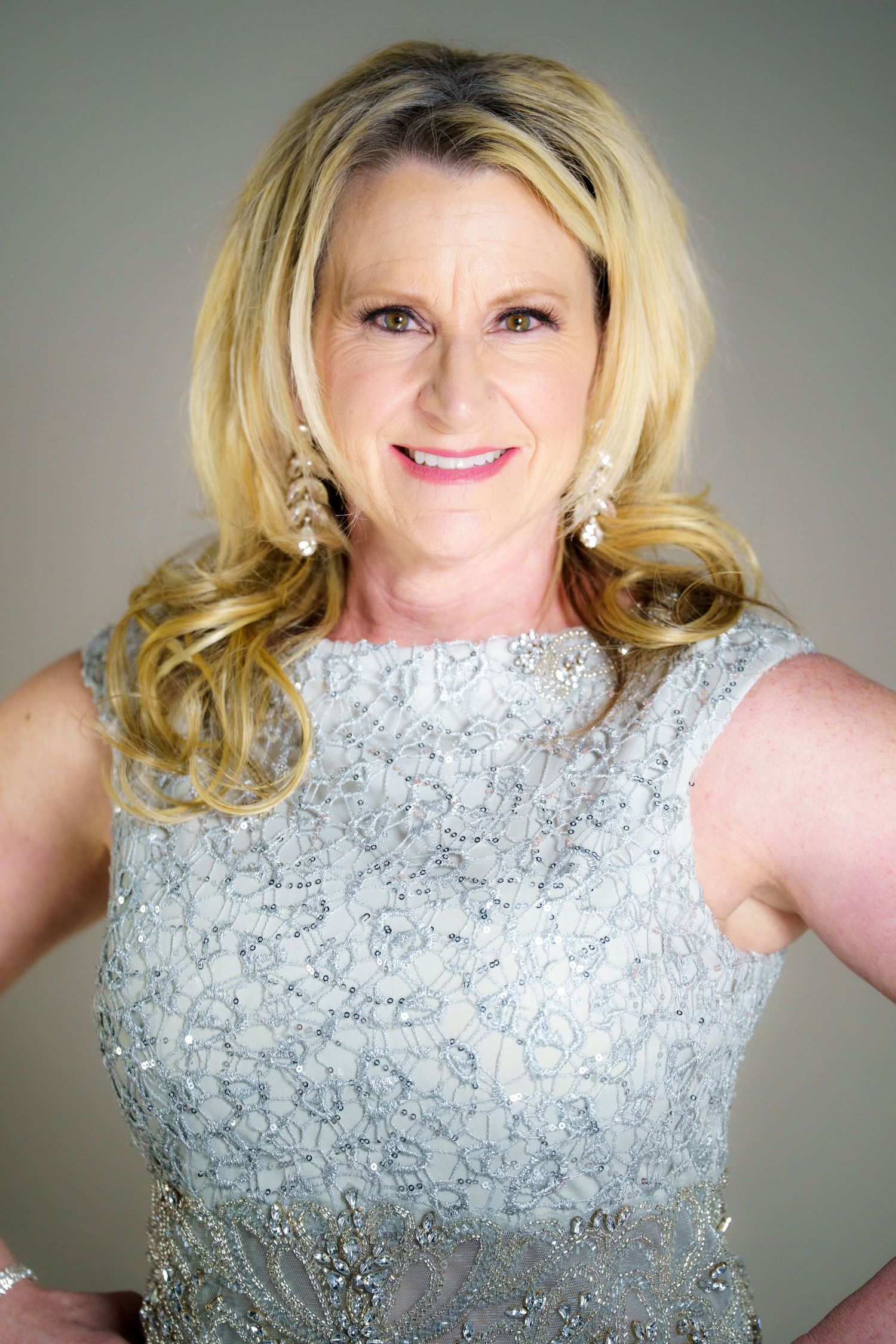A blonde wearing a sleeveless silver lace dress and pearl earrings poses against a gray studio backdrop.