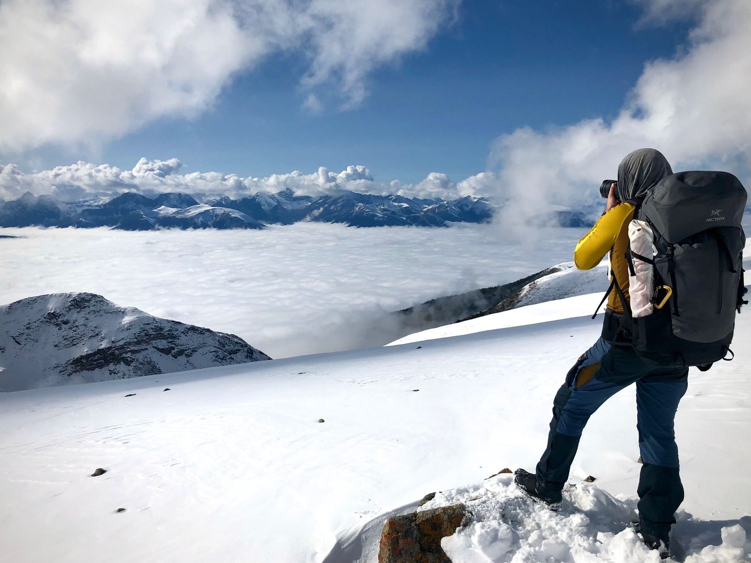 Adventurous hiker with large backpack stands on snowy mountain peak overlooking cloud-covered valley and mountain range in Jasper