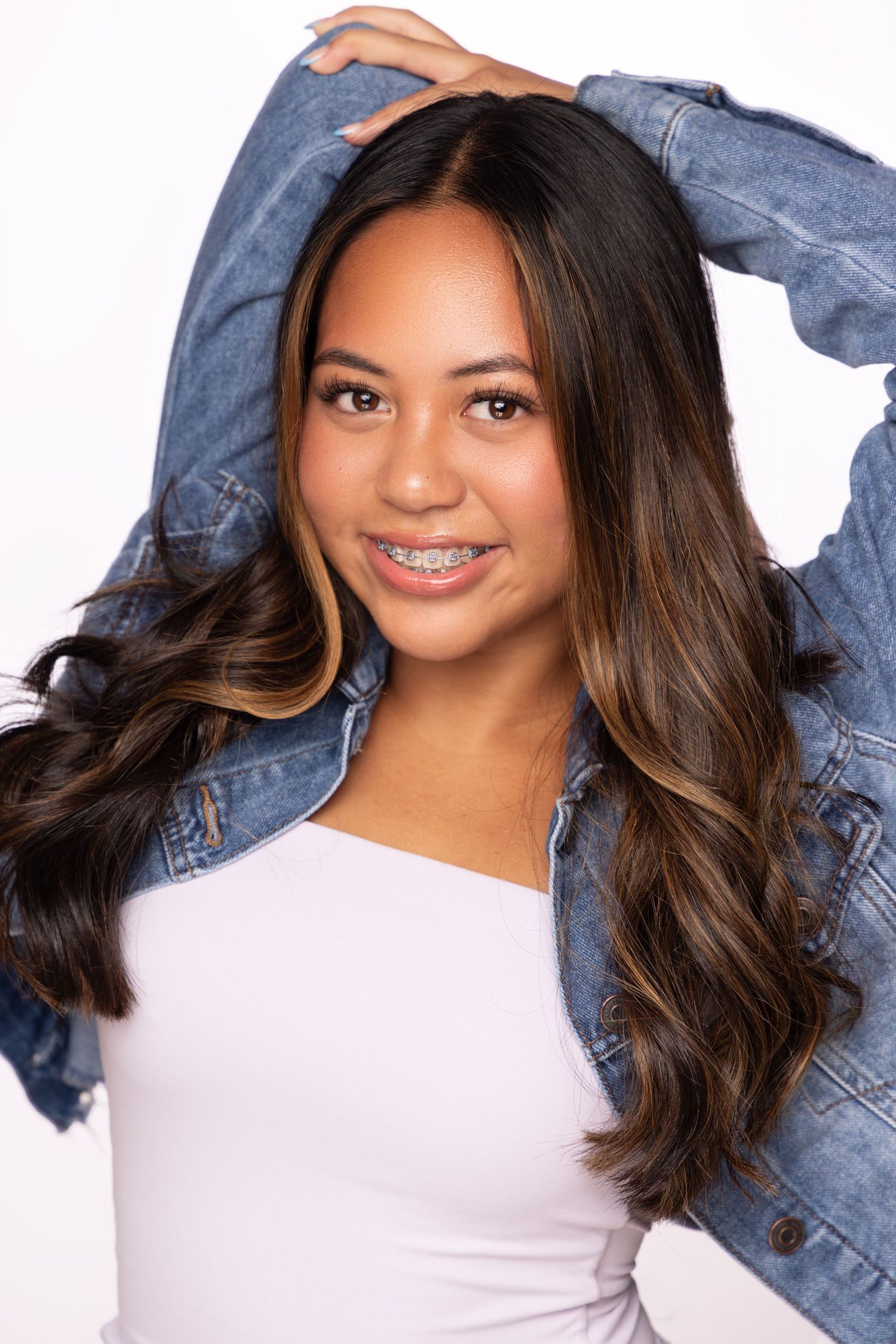 Woman with long wavy brown hair wearing white top and denim jacket poses against white background with bright smile.