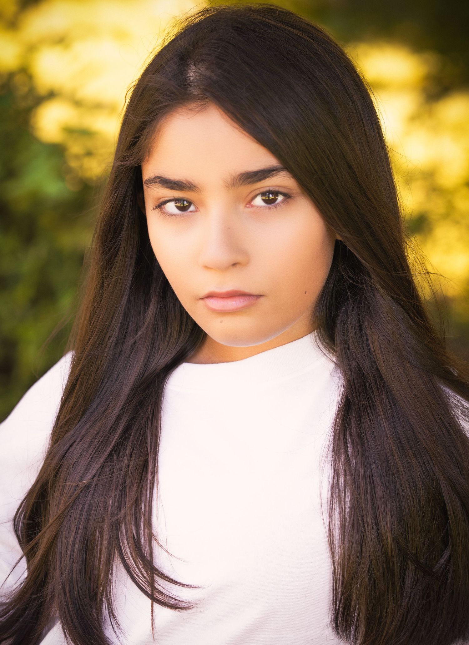Portrait photo with long dark hair against blurred yellow nature background wearing white top.