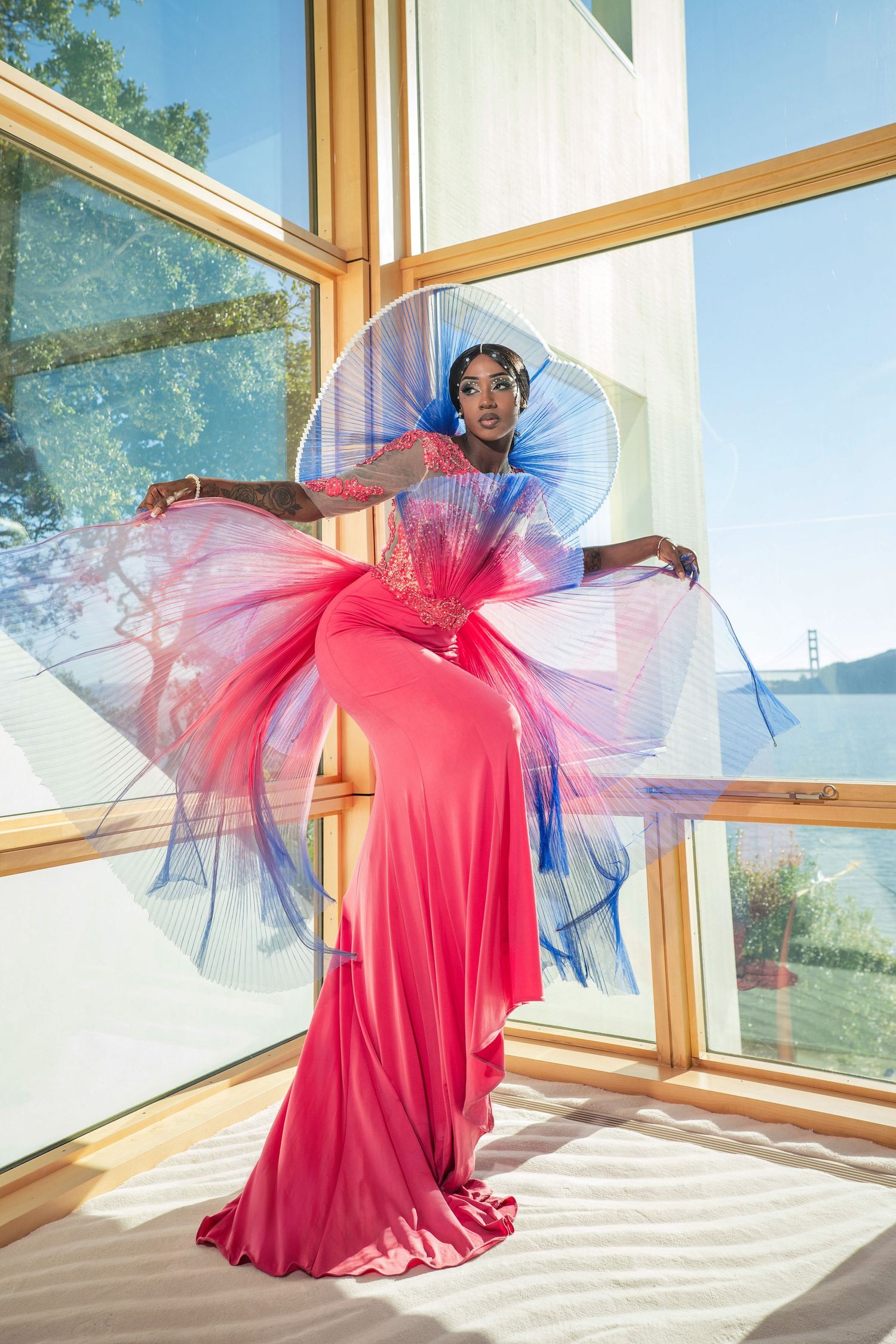 A dramatic fashion pose in a flowing pink and blue tulle gown by large windows in natural sunlight.