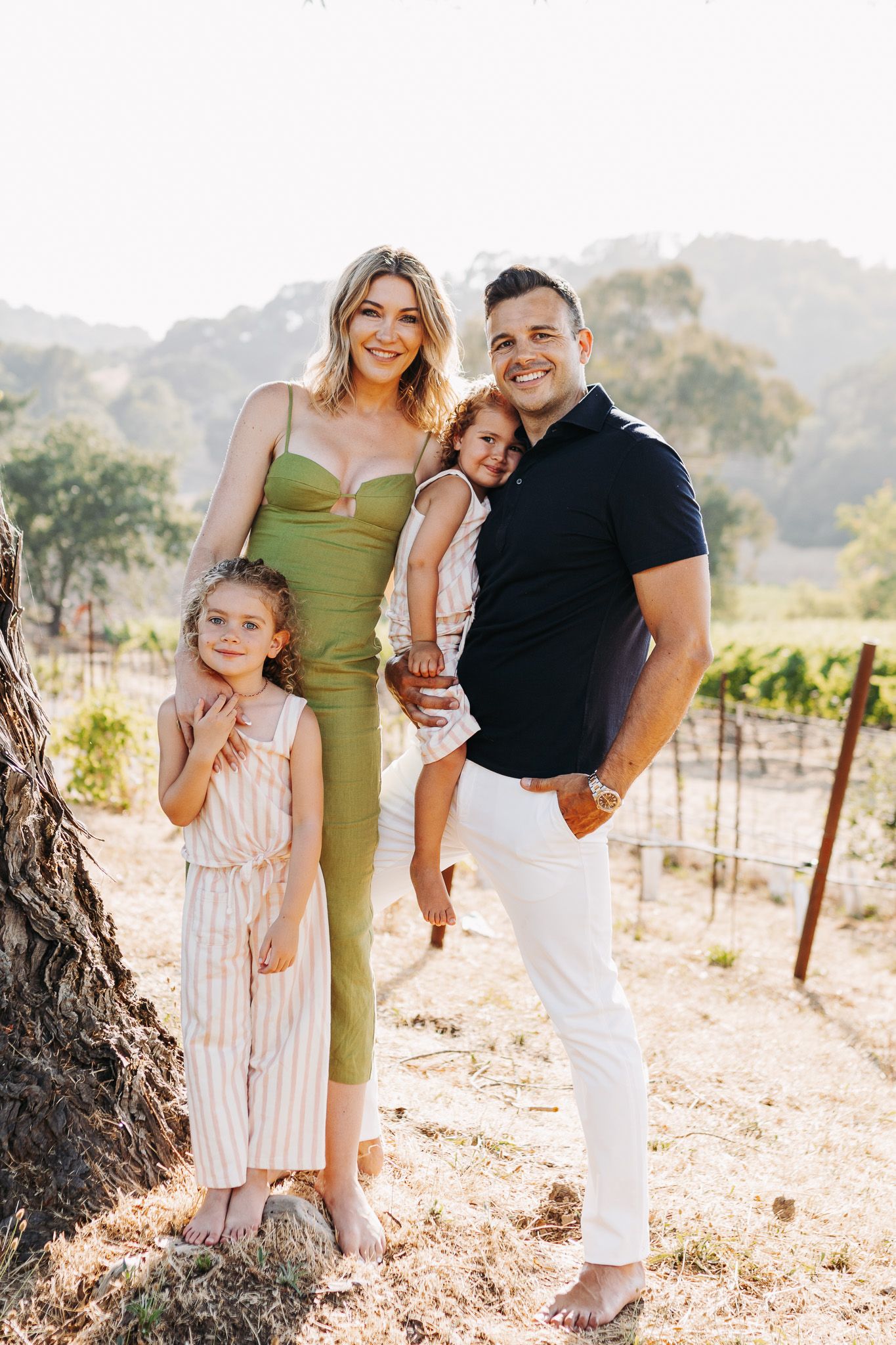 A young family poses together on a sunny day in a vineyard against mountainous backdrop.