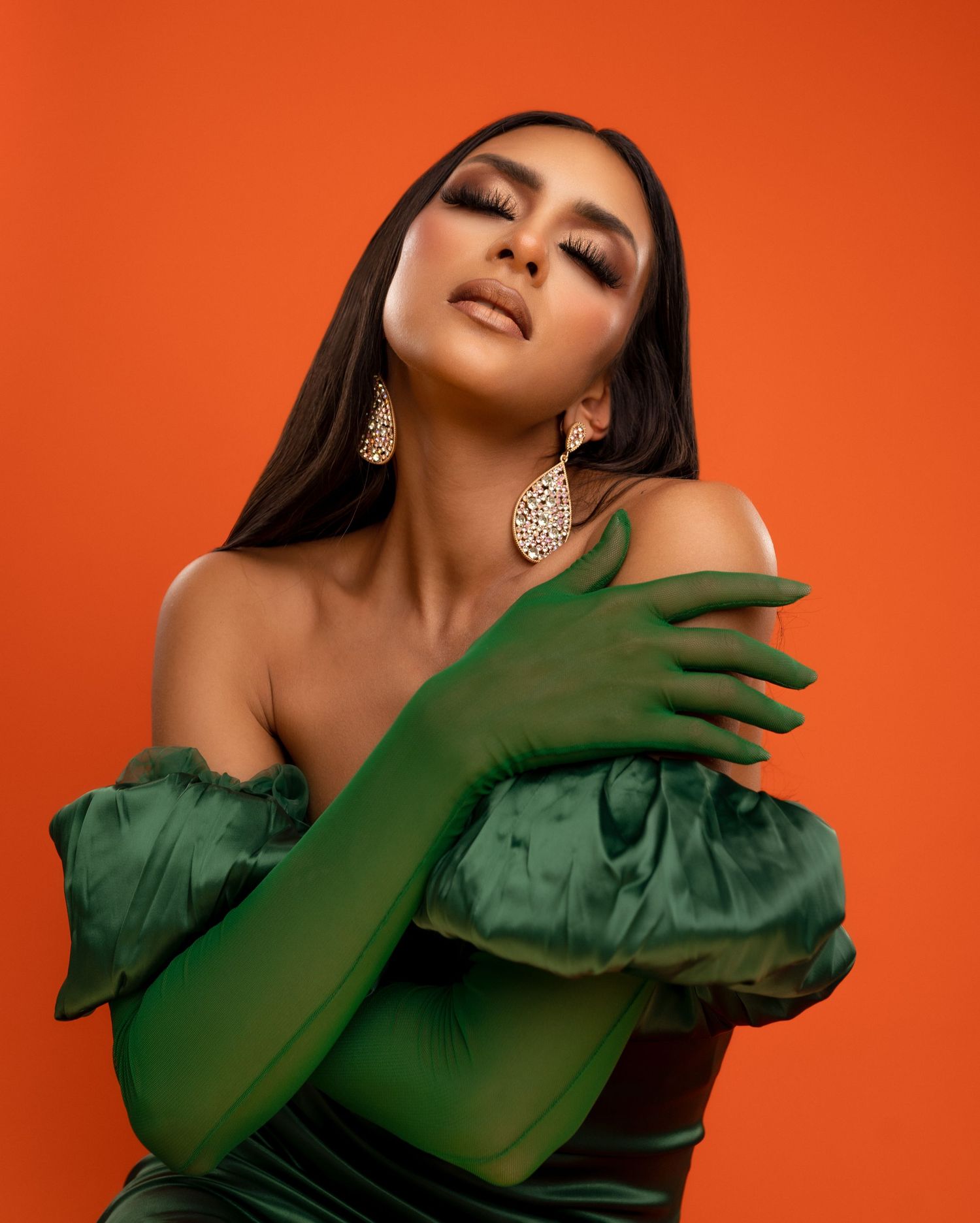 Glamorous model in emerald green gloves and gown poses against vibrant orange background with statement earrings.