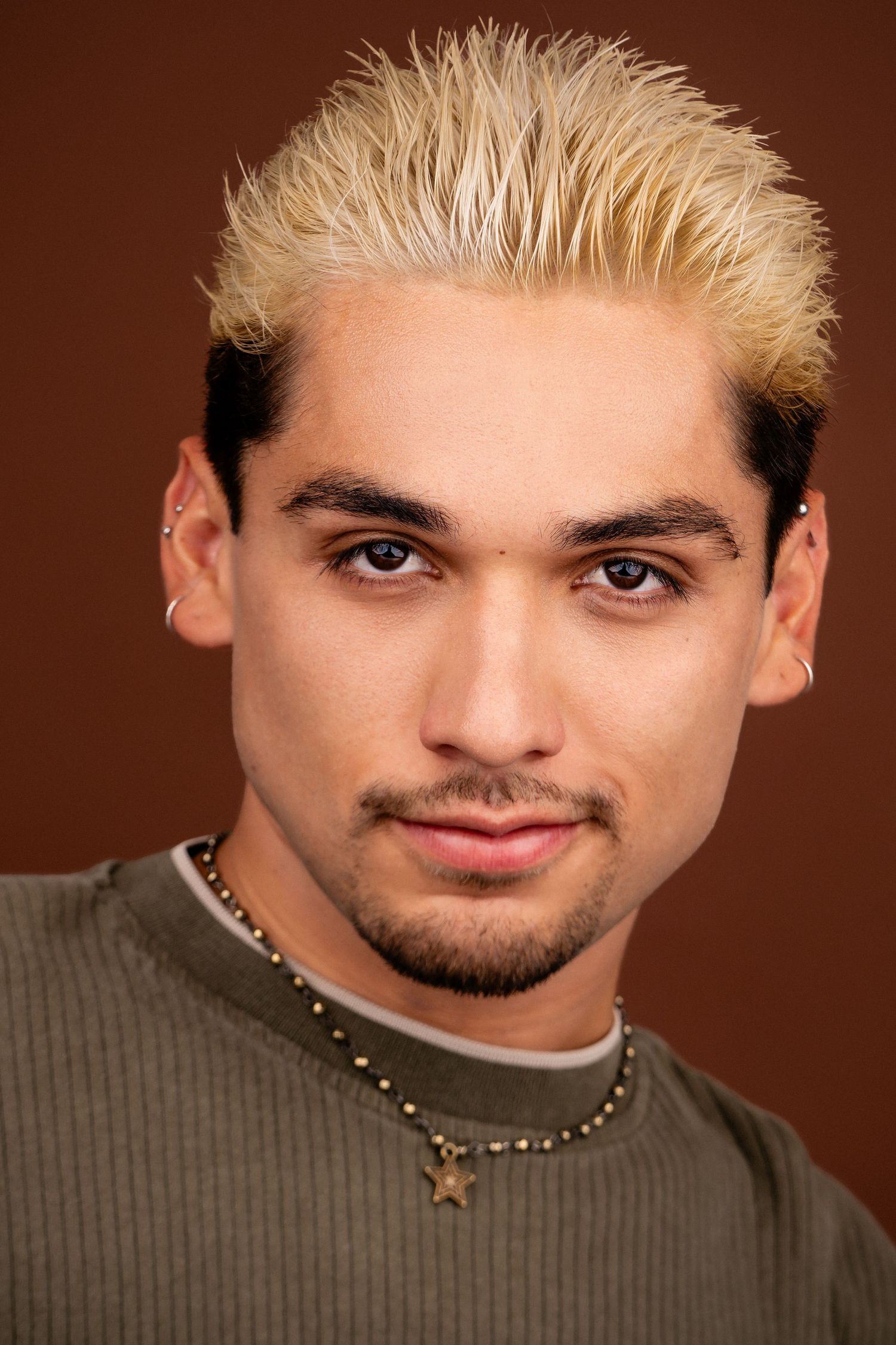 Portrait shot of someone with bleached blonde spiked hair wearing a brown ribbed shirt and star pendant necklace.