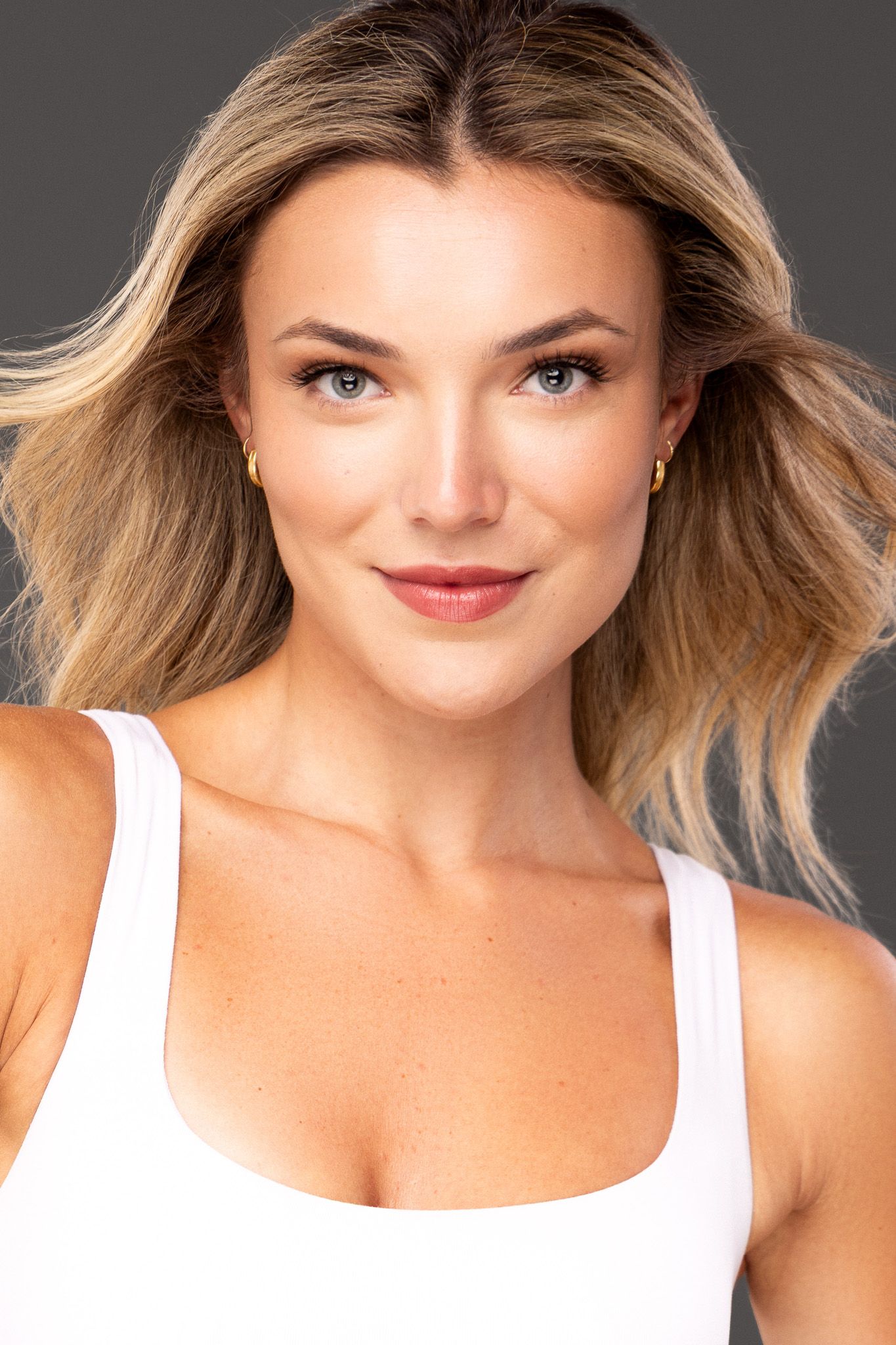 Professional studio portrait of a smiling model in a white tank top with flowing blonde hair against a gray background.