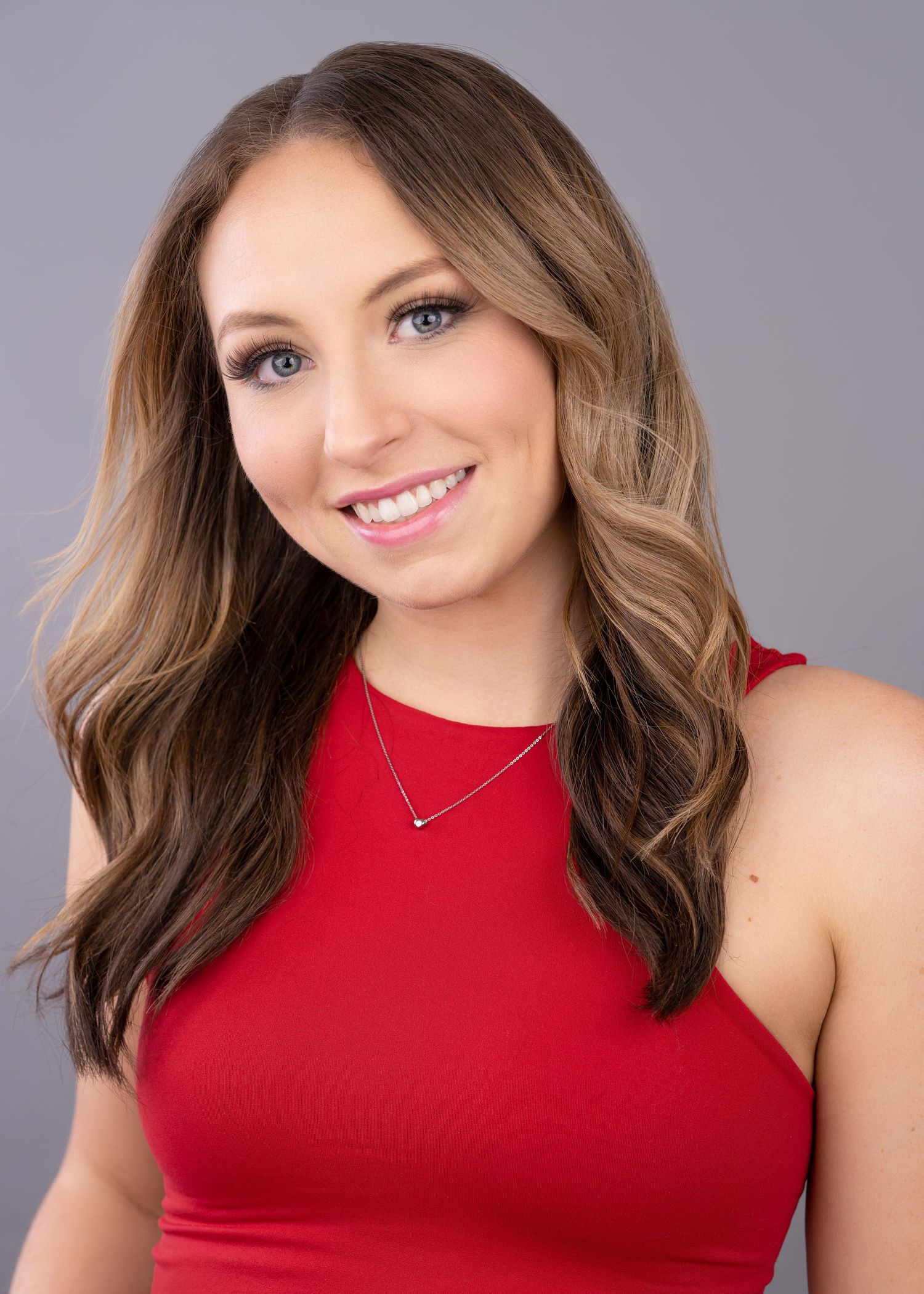 Portrait of a smiling person with wavy brown hair wearing a red sleeveless top and delicate necklace.