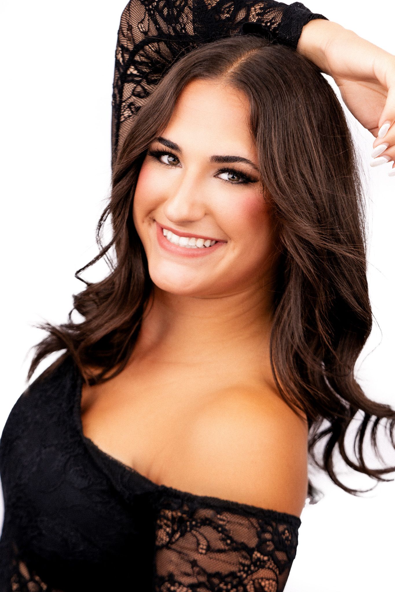 Smiling woman with long dark wavy hair wearing black lace off-shoulder top against white background.