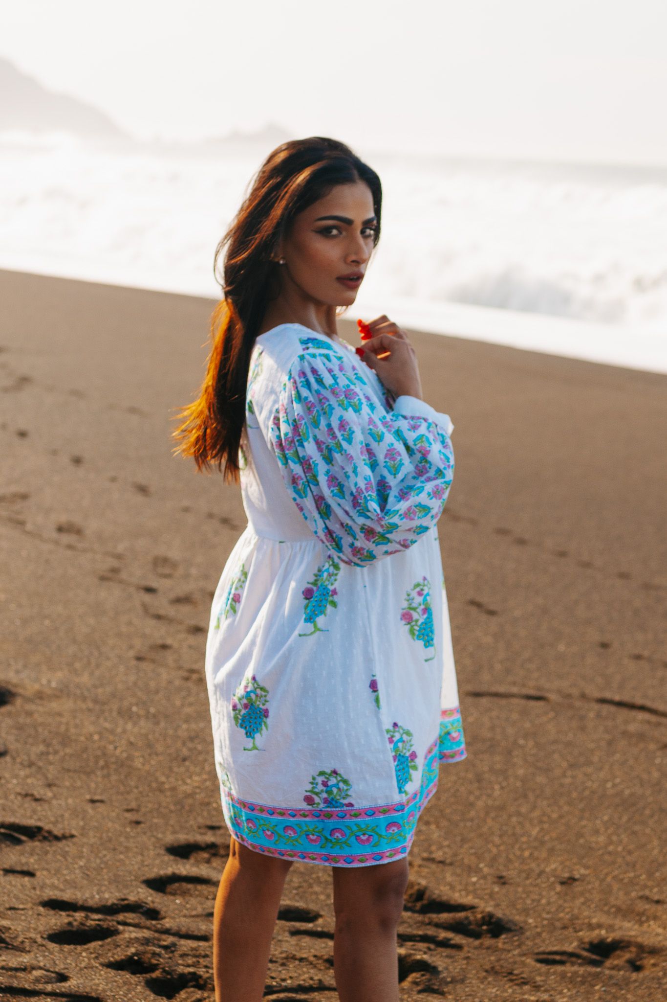 A person in a white and turquoise floral dress poses on a sandy beach during sunset with windswept hair.