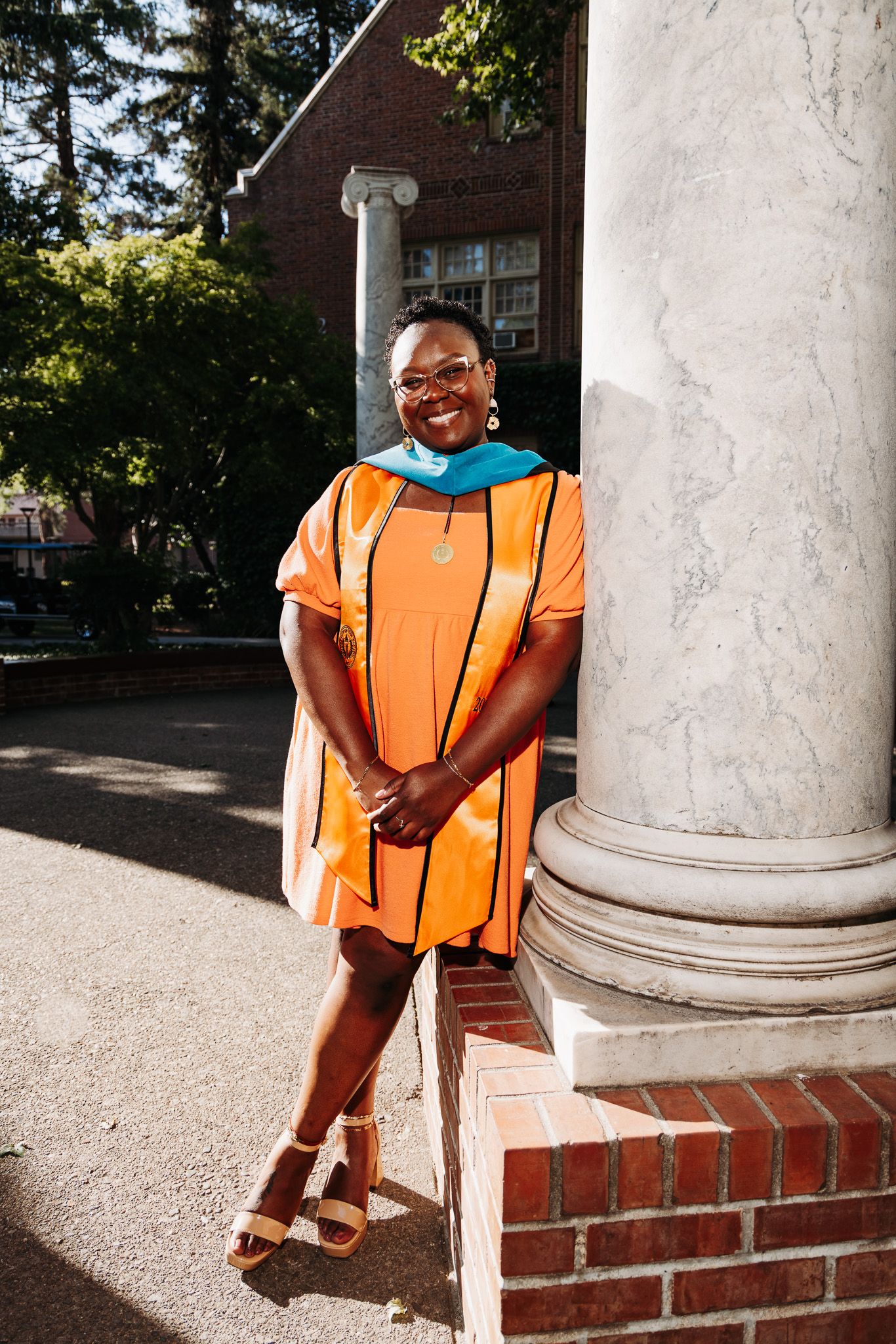 A graduate in orange academic regalia stands smiling against a white column and brick building on a sunny day.