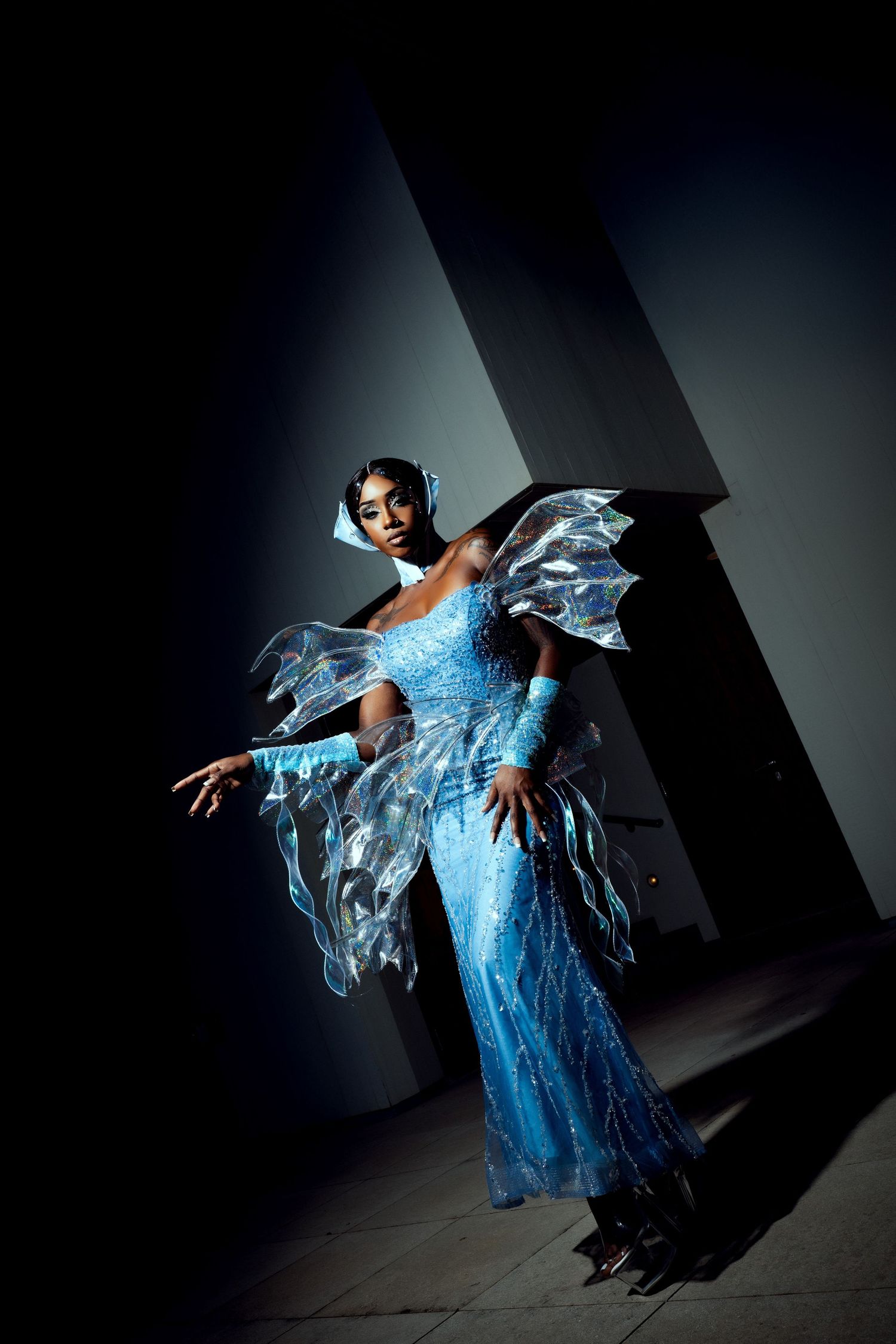 A performer in a flowing blue crystalline dress with wing-like elements poses dramatically under theatrical lighting.