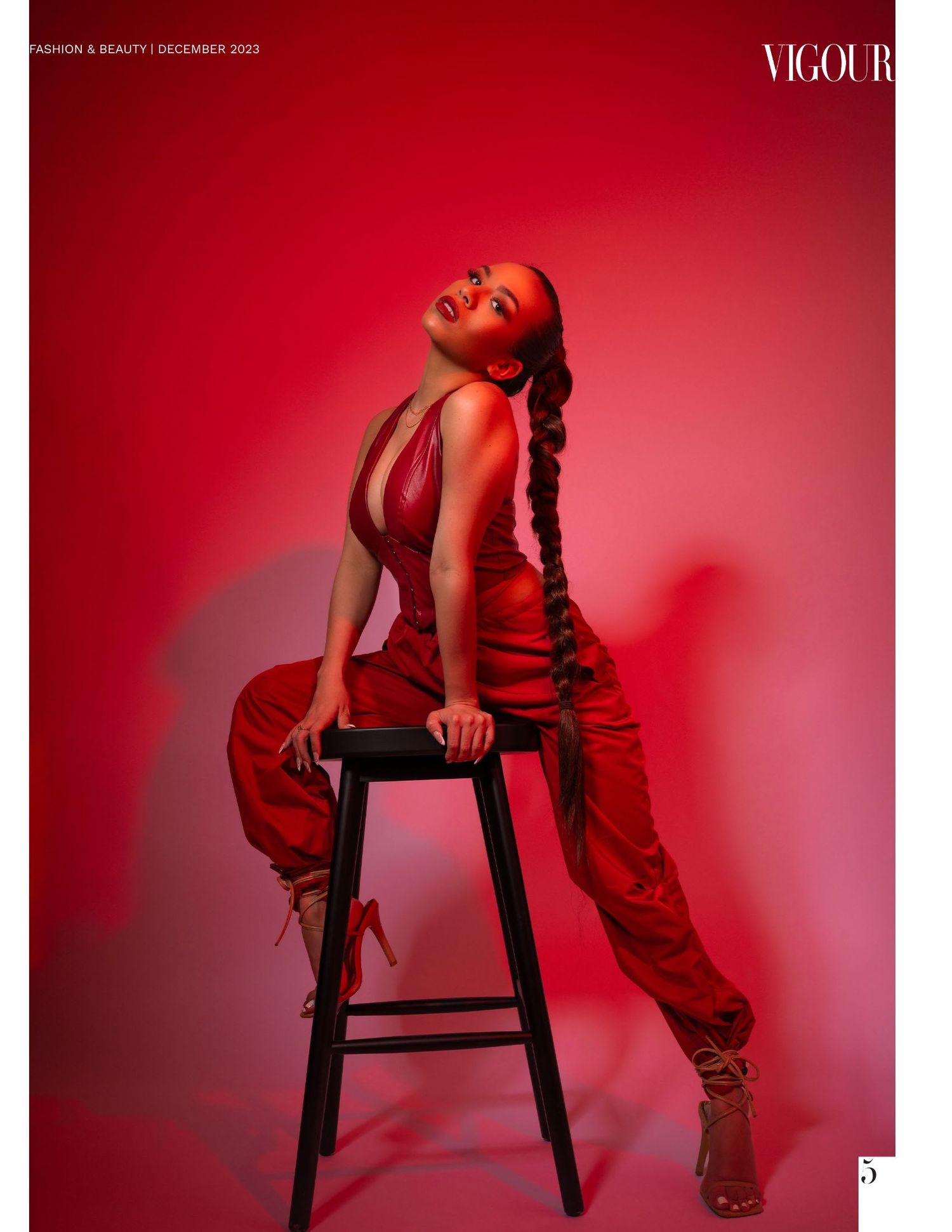 Model poses dramatically on stool wearing red outfit against vibrant crimson backdrop with dramatic lighting.