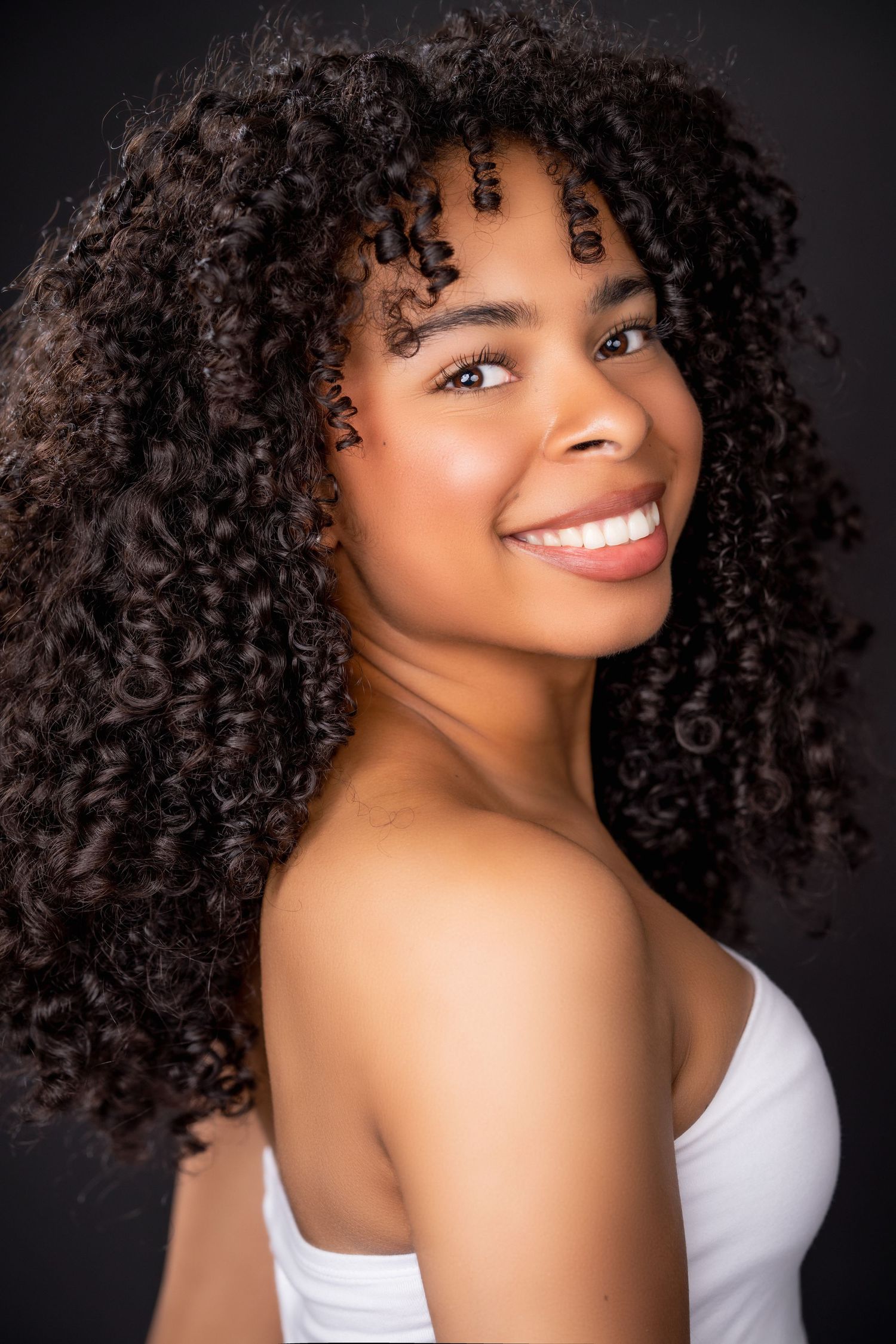 Beauty portrait of a smiling model with voluminous natural curly hair wearing a white tank top against dark background.