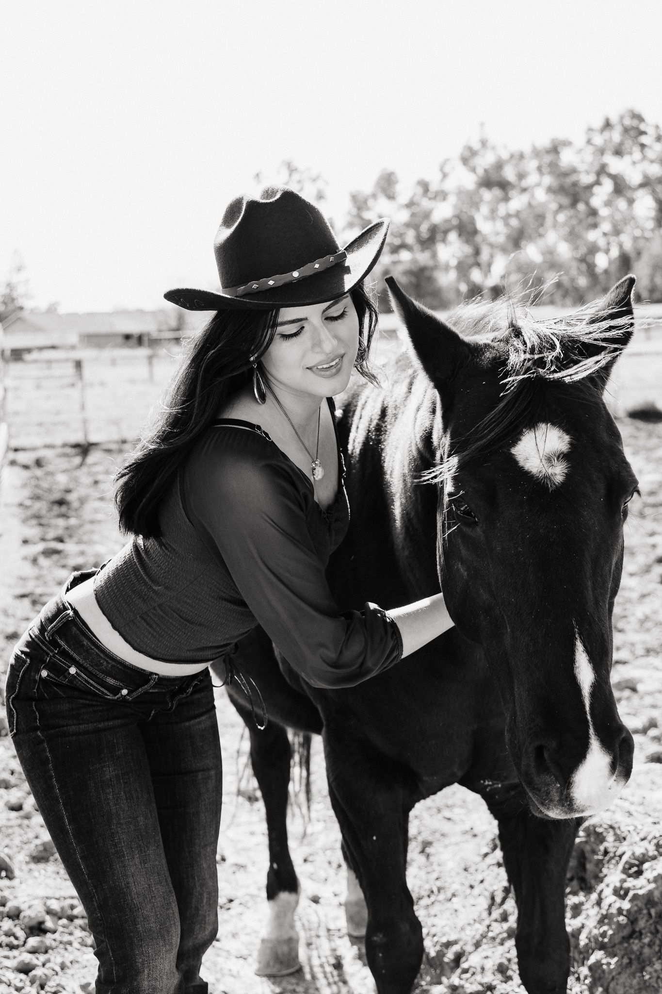 Black and white photo of a cowgirl in western attire sharing an affectionate moment with a dark horse outdoors.